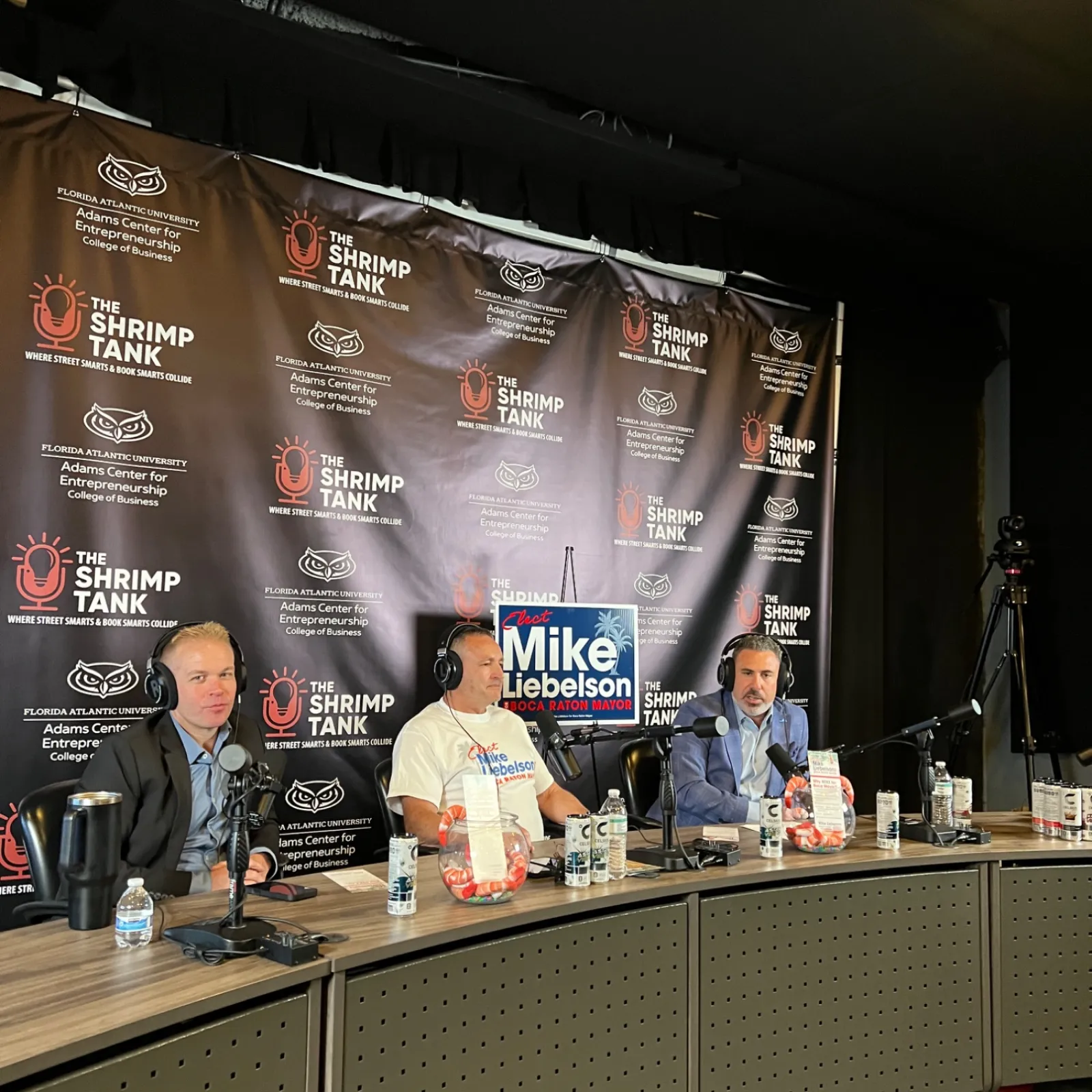 Three men recording a podcast at The Shrimp Tank with microphones, headphones, and a campaign sign for Mike Rodelson.