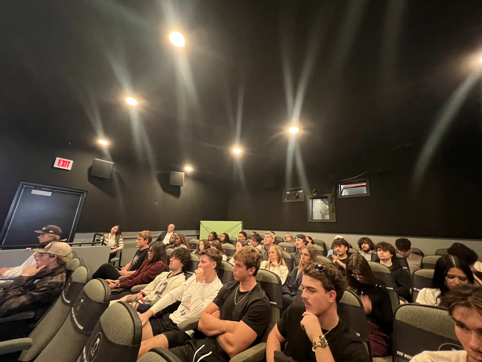 Audience seated and attentively watching in a small, dark theater with overhead spotlights.