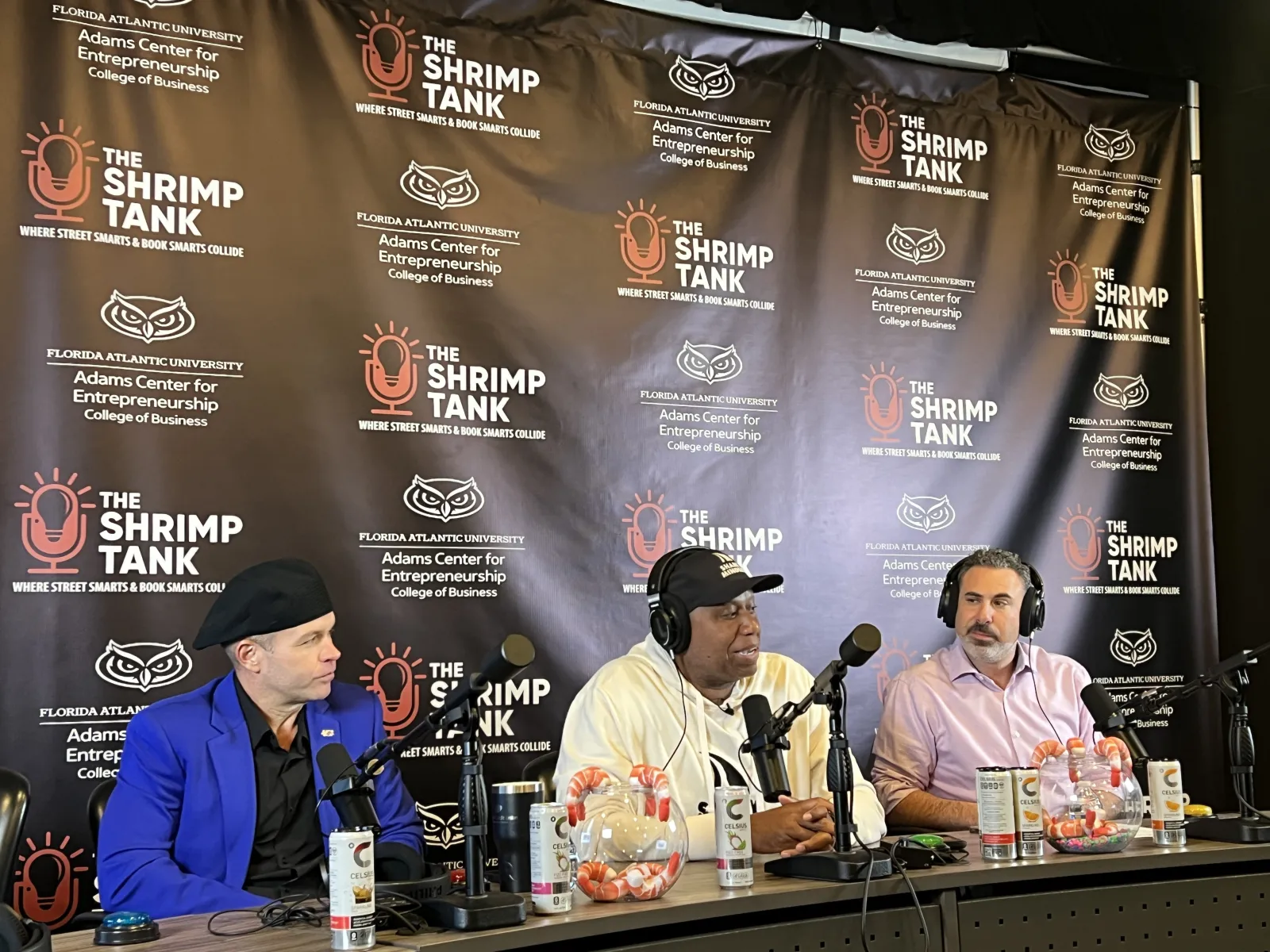 Three men speaking into microphones during The Shrimp Tank podcast with branded backdrop and shrimp bowls.