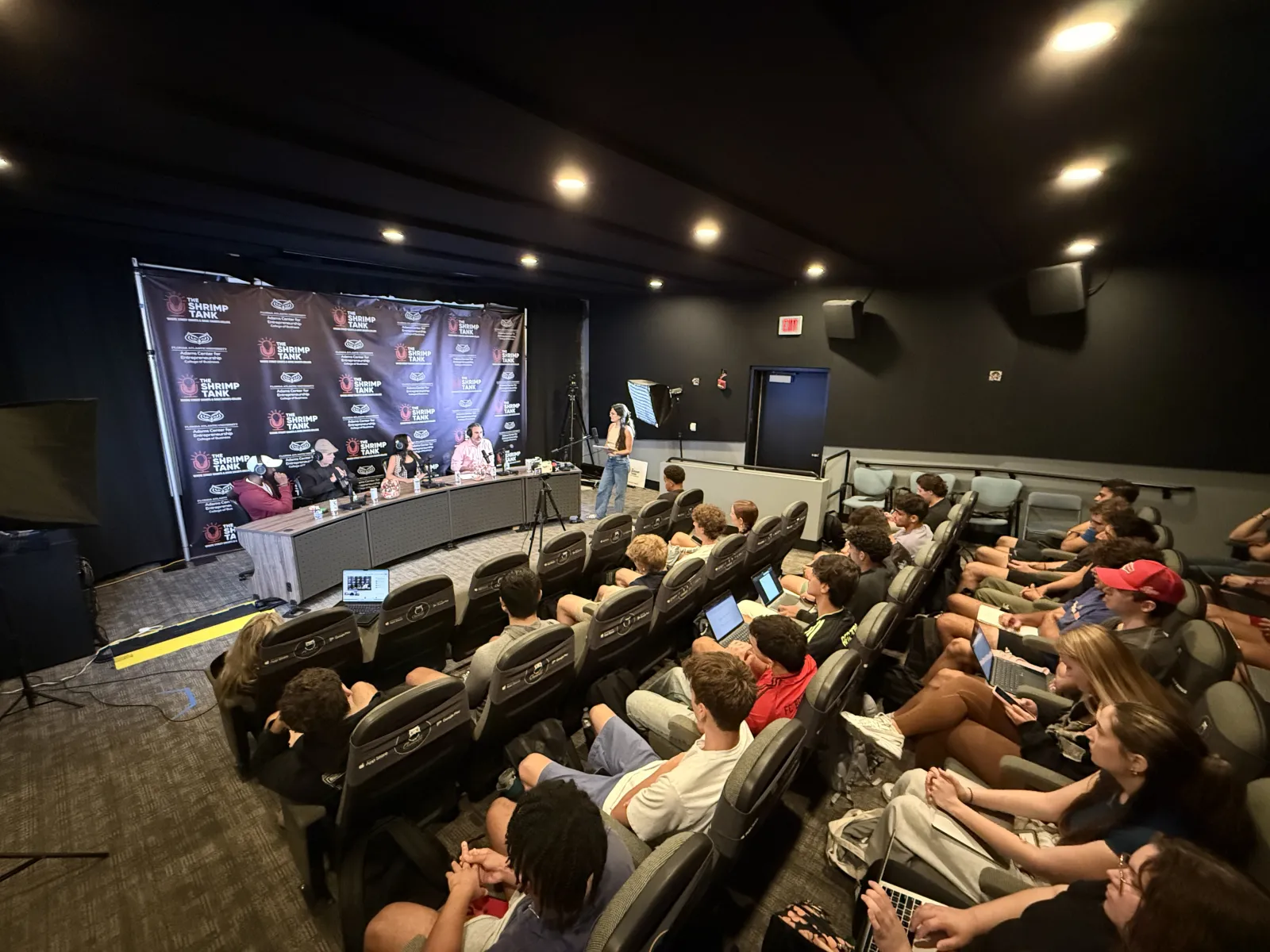 Conference room with panel discussion, attendees in theater seats using laptops and cameras recording event