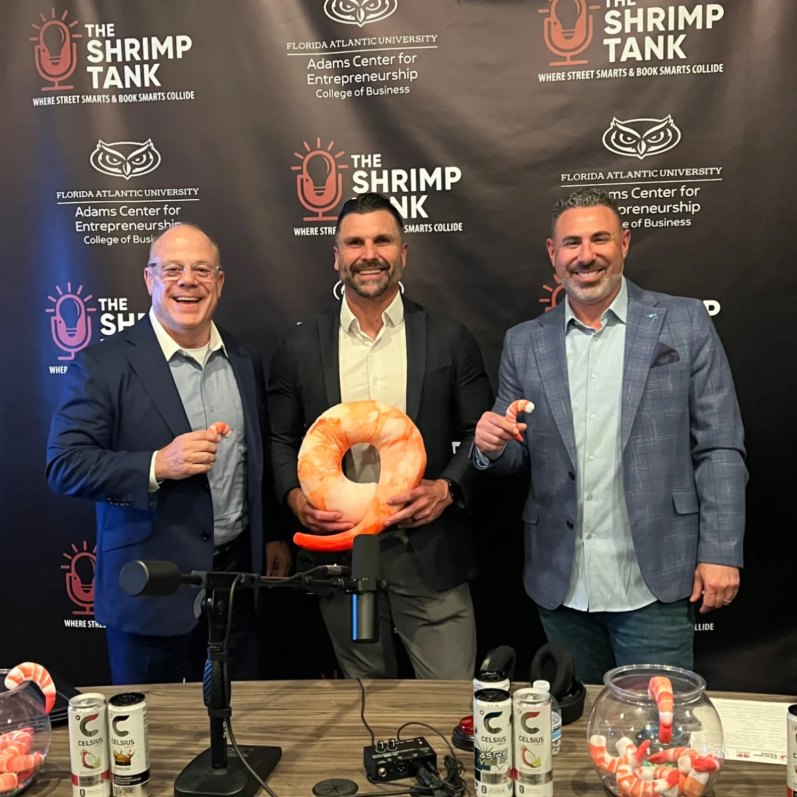Three men in suits smiling at a Shrimp Tank event with shrimp-shaped props and beverages on a table.