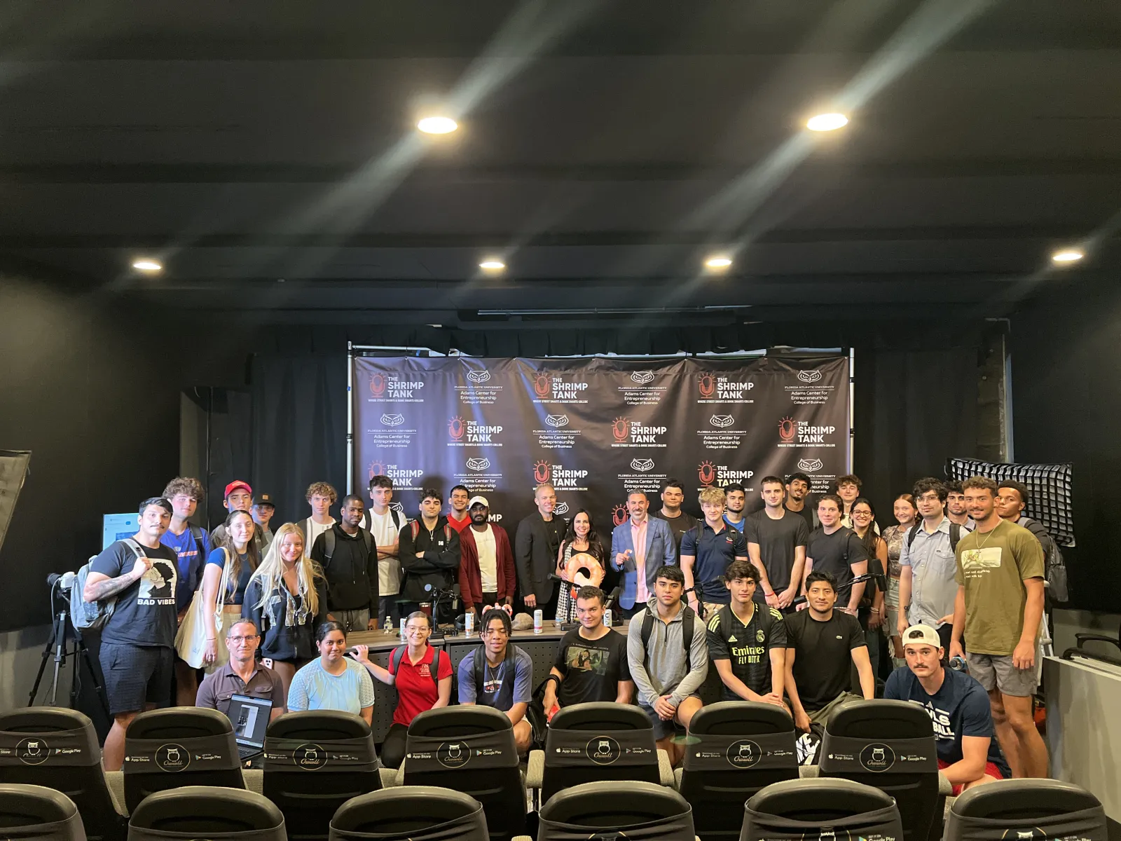 Large group of diverse young adults and professionals posing on stage in front of a Shrimp Tank event banner