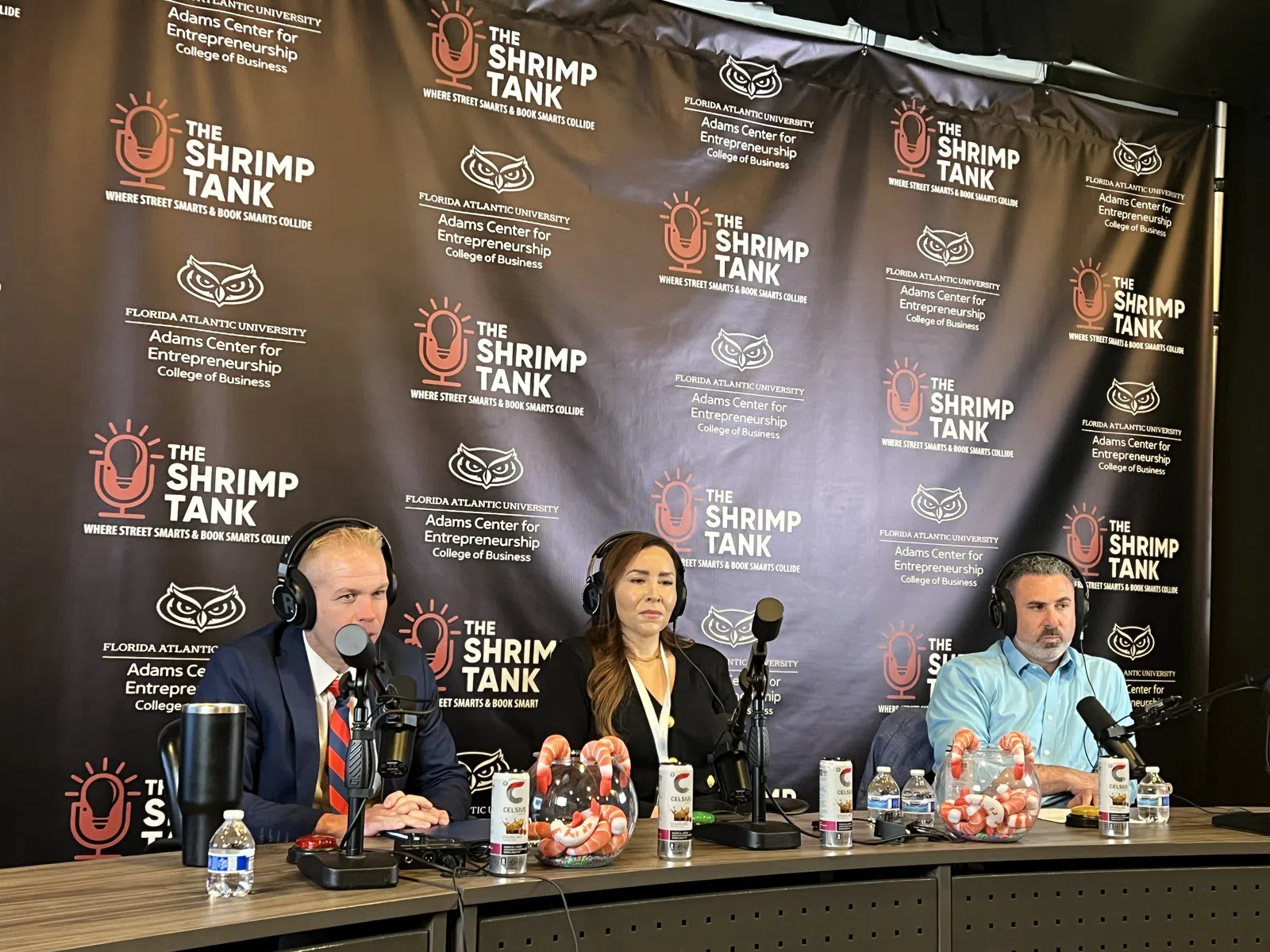 Three hosts speaking into microphones on a podcast set with The Shrimp Tank backdrop and shrimp decor on table.
