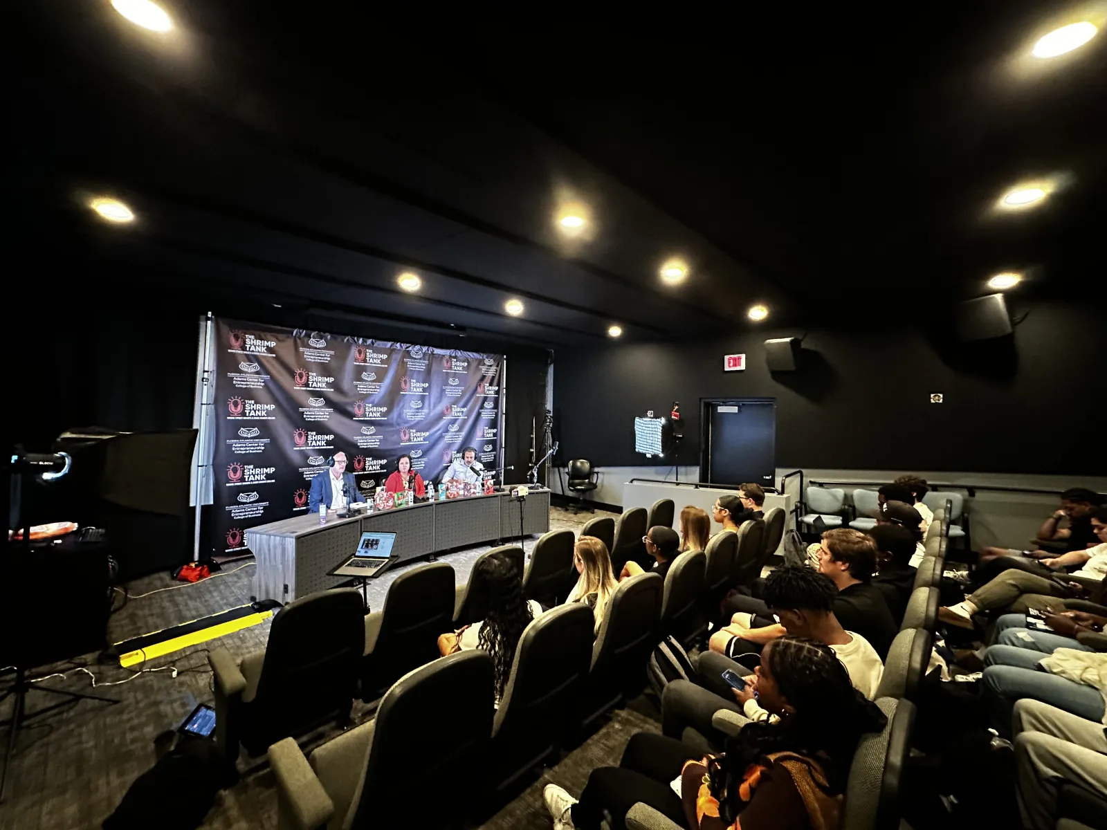 a group of people sitting in a room with a stage and a large screen