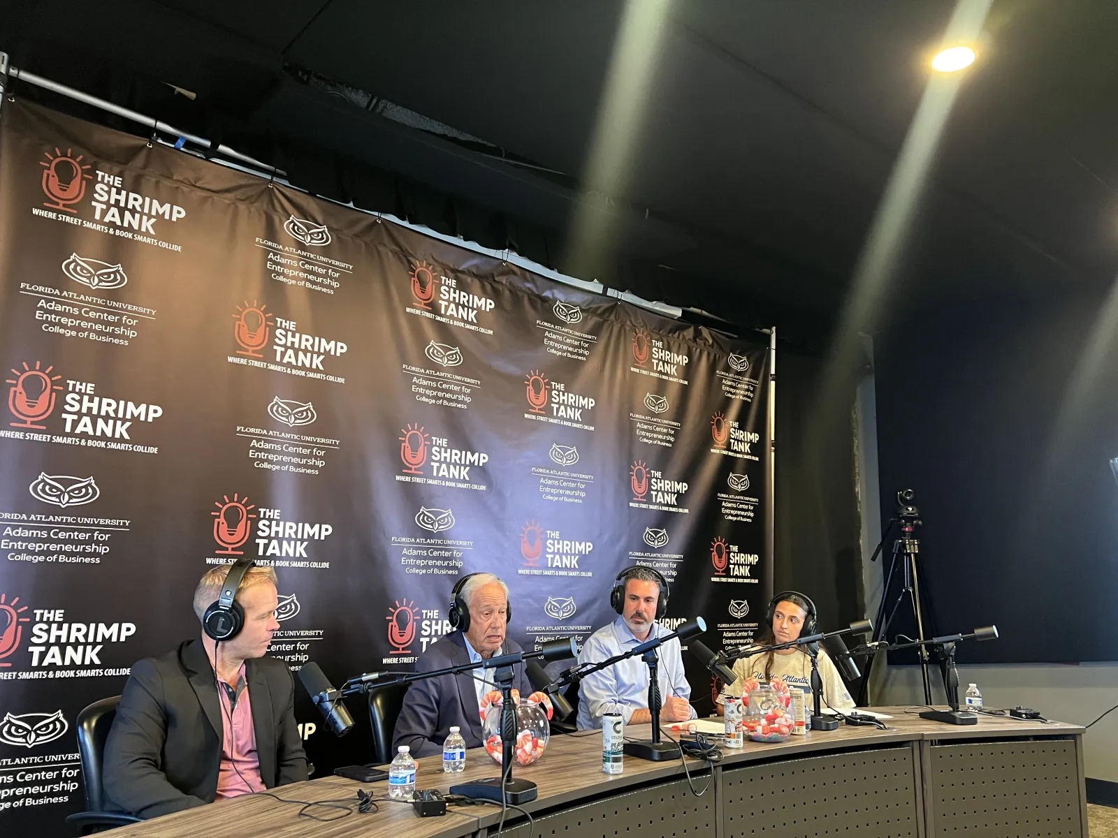 Panel of four speakers with microphones in recording studio for The Shrimp Tank podcast with branded backdrop and lighting.