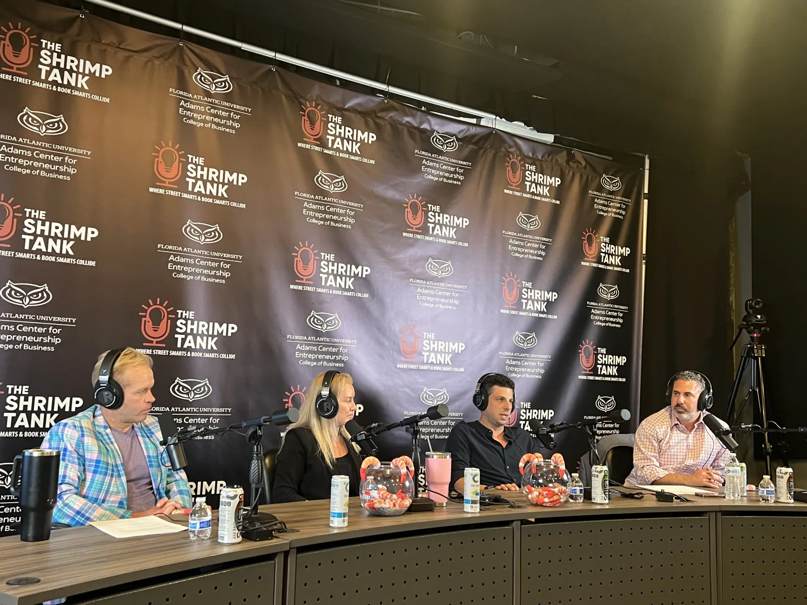 Four hosts wearing headphones speaking into microphones at The Shrimp Tank podcast panel with branded backdrop