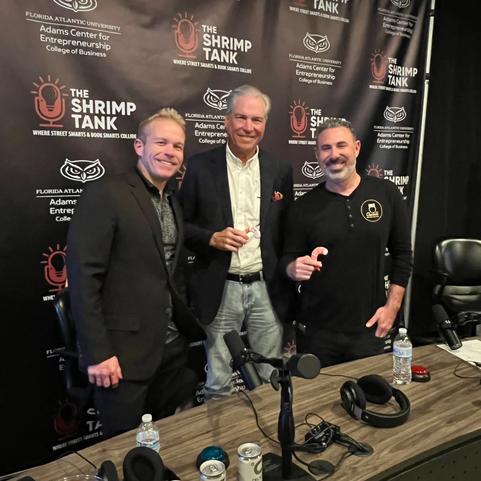Three men posing in front of The Shrimp Tank podcast backdrop with a table of podcast equipment and drinks.