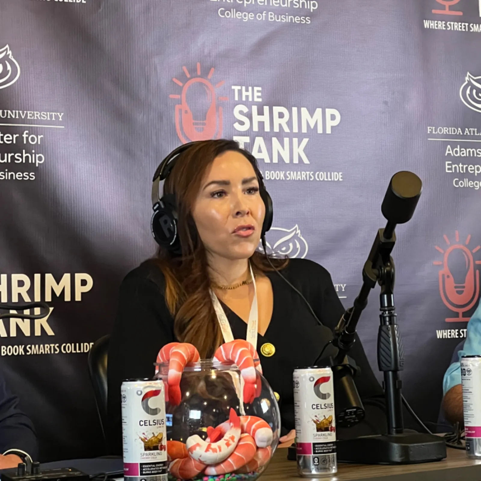 Woman wearing headphones speaks into microphone at The Shrimp Tank event with promotional beverages displayed.