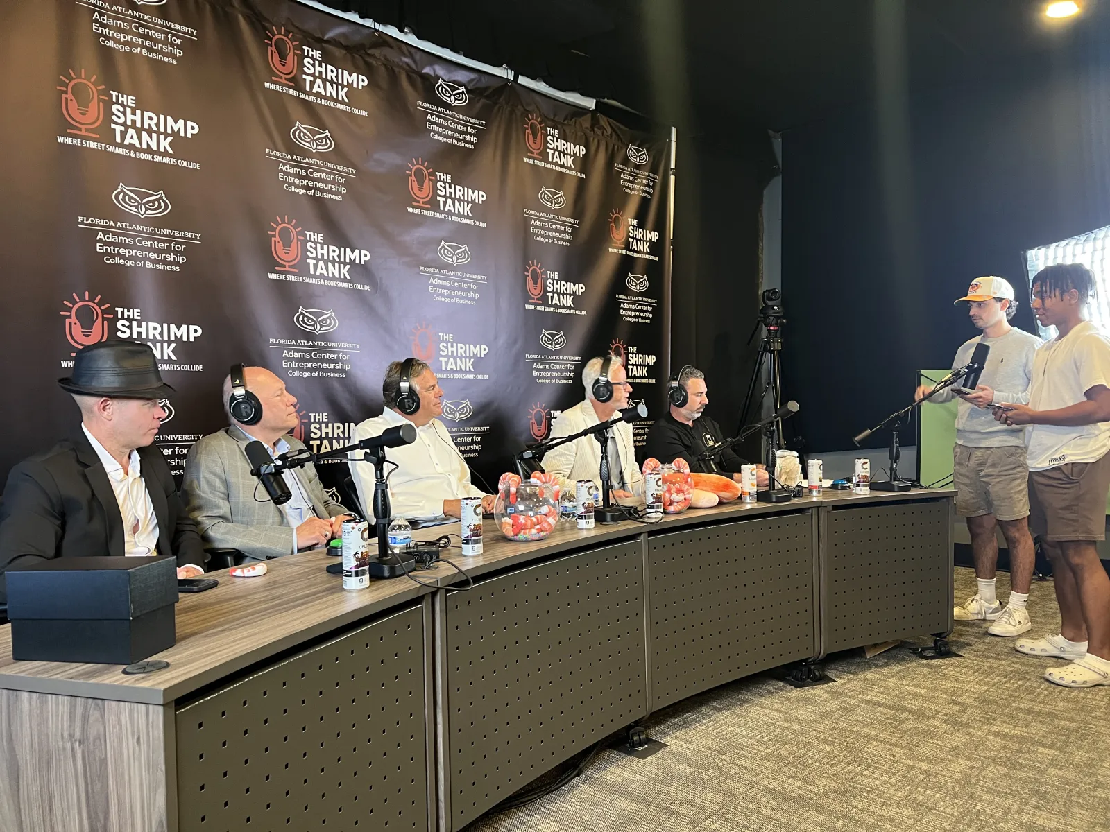 Panel of five men recording a podcast at The Shrimp Tank event with two men hosting on the side.
