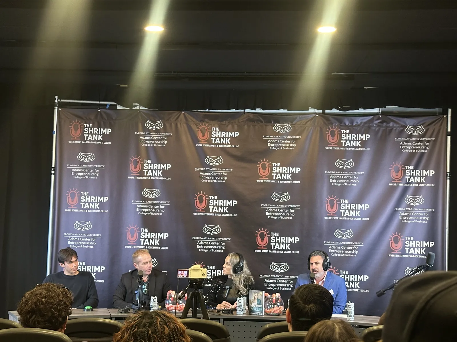 Panel of four speakers at The Shrimp Tank event with branded backdrop and audience in foreground