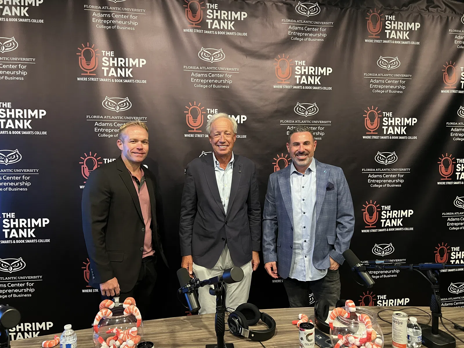 Three men standing in front of The Shrimp Tank podcast backdrop with microphones and headphones on table