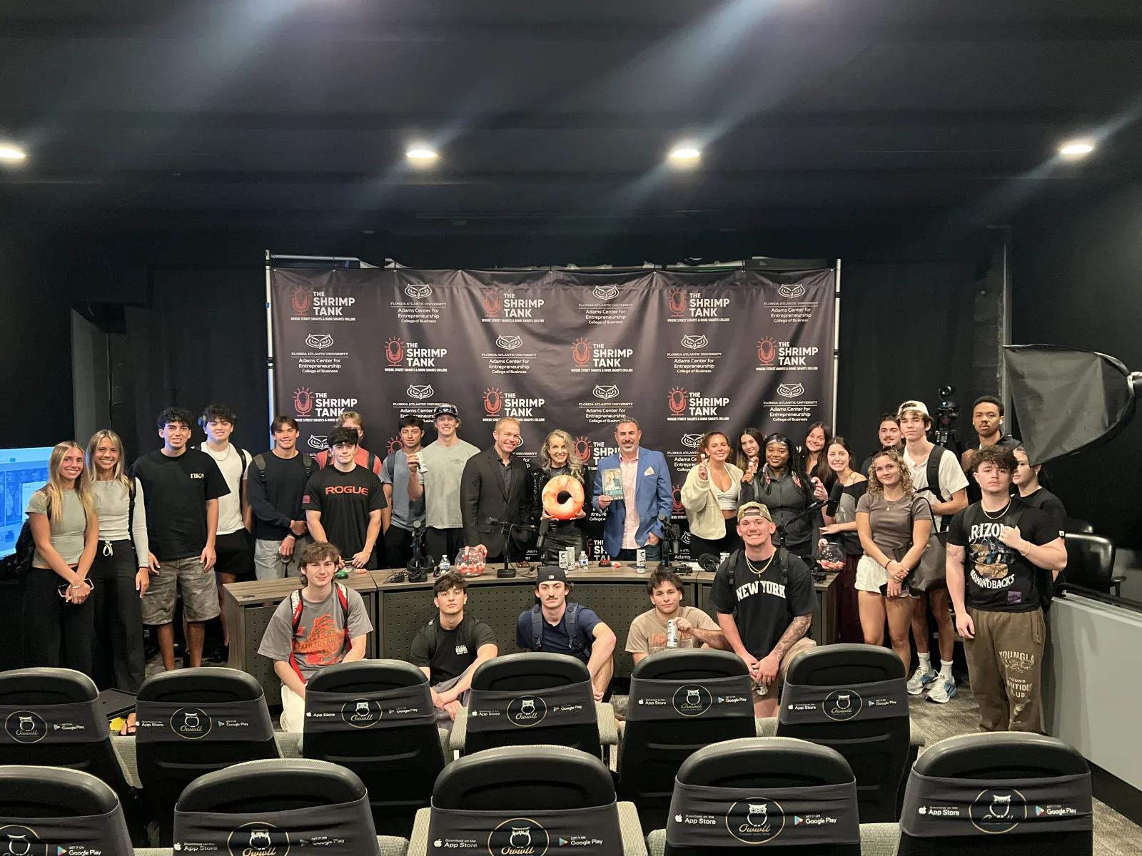 Large group of diverse people posing in front of The Shrimp Tank backdrop in a studio setting with chairs in foreground.