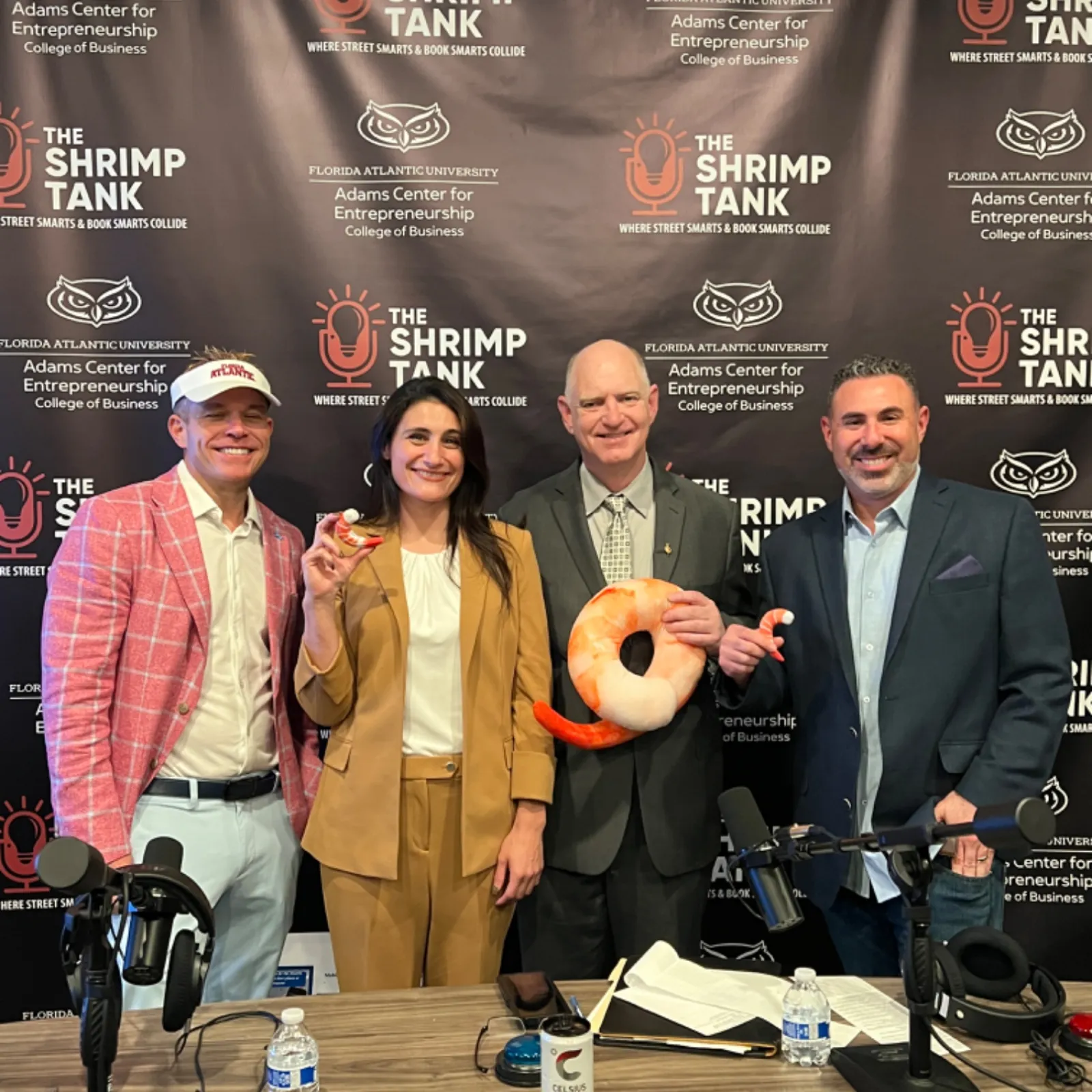Four people smiling at a shrimp-themed podcast with microphones and branded backdrop for The Shrimp Tank podcast.