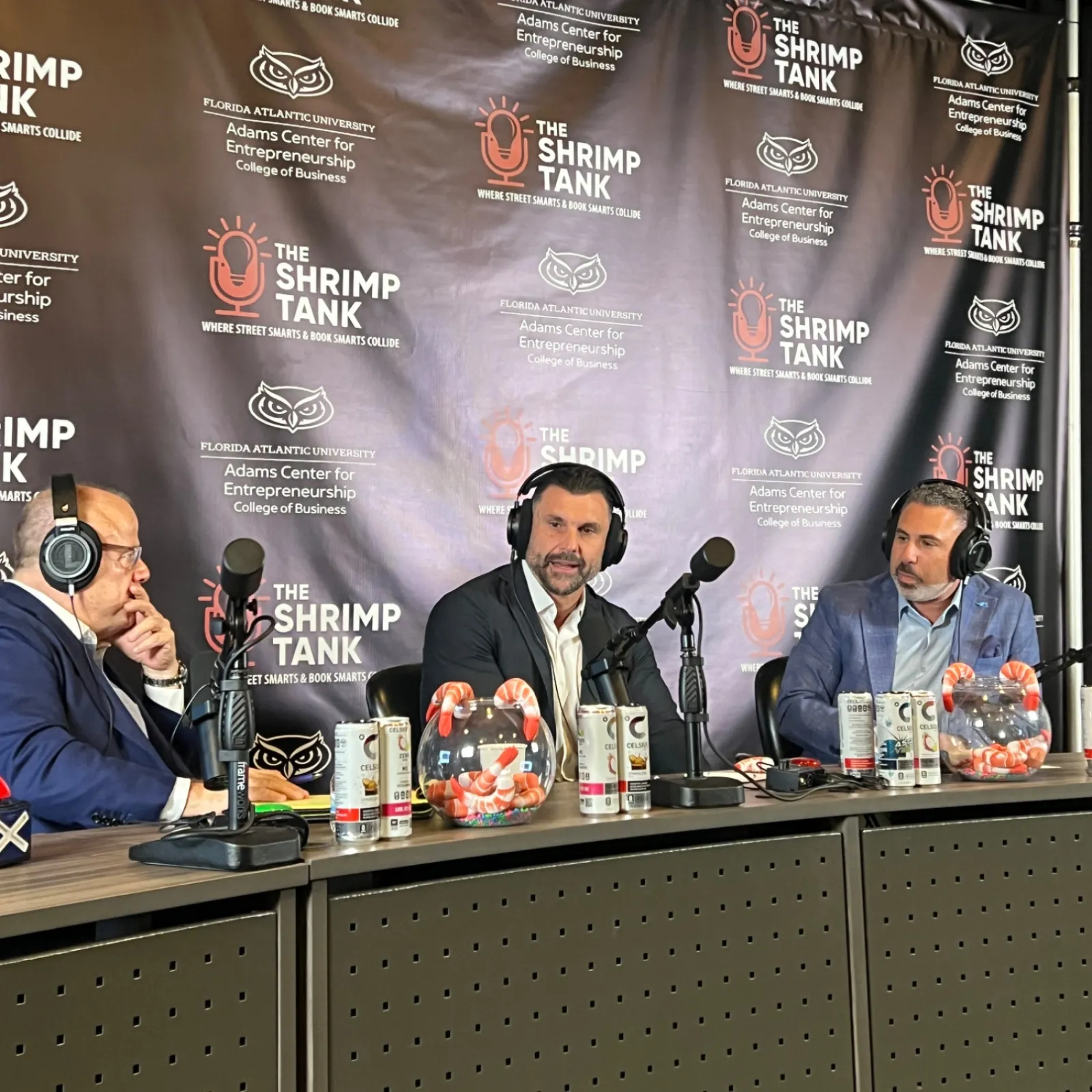 Three men wearing headphones sitting at a podcast table with microphones and shrimp tanks beside them.