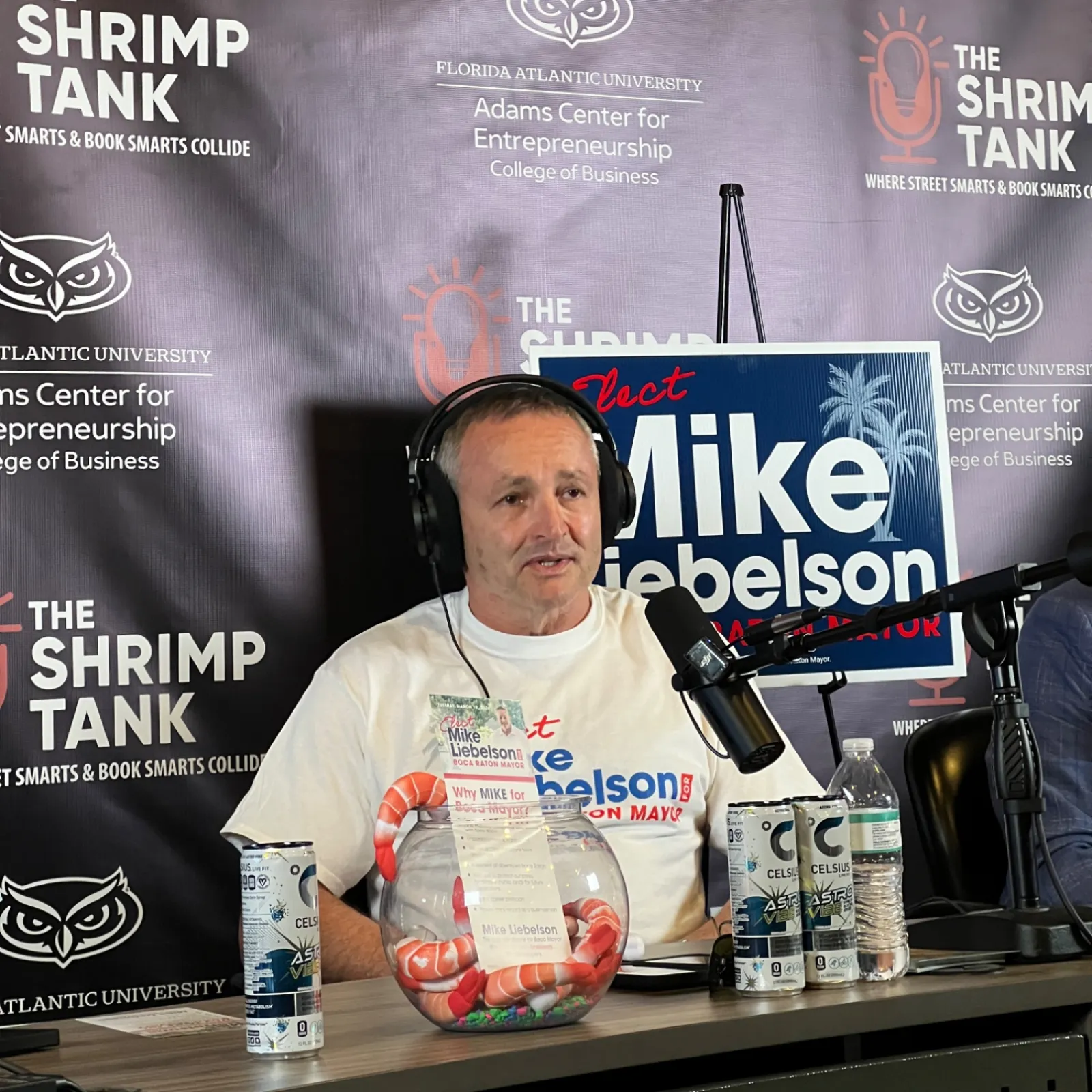 Man wearing headphones speaks into microphone on podcast set with shrimp tank backdrop and campaign sign.