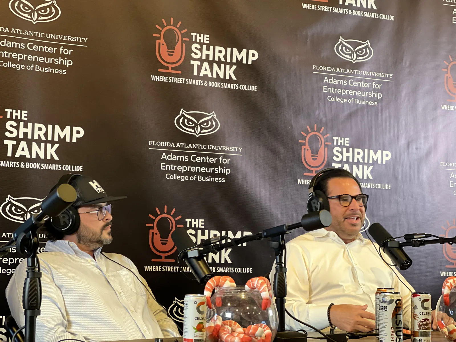 Two men wearing headsets speaking into microphones during a podcast at The Shrimp Tank in front of a branded backdrop.