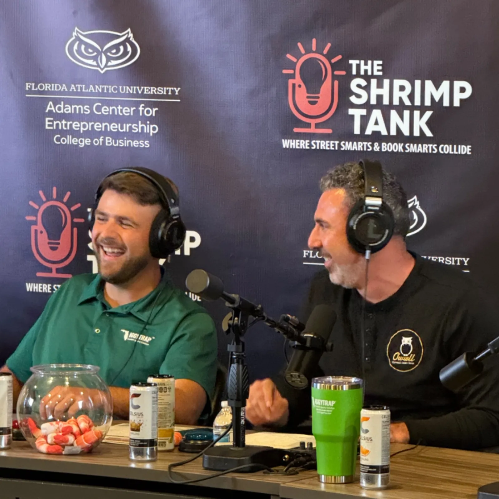 Two men wearing headphones recording a podcast at a table with microphones and drinks in front of a Shrimp Tank backdrop.
