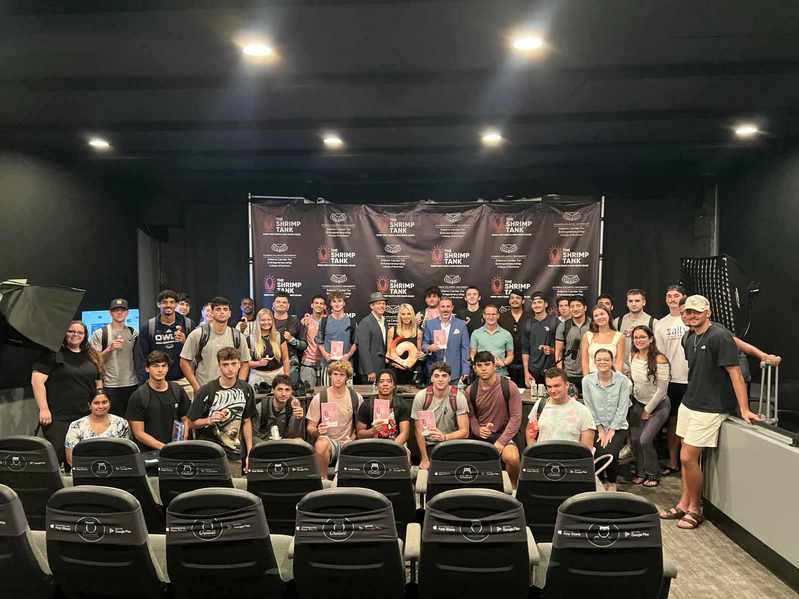 Large group of young adults posing in a small theater with a Shrimp Tank branded backdrop and seats in foreground.