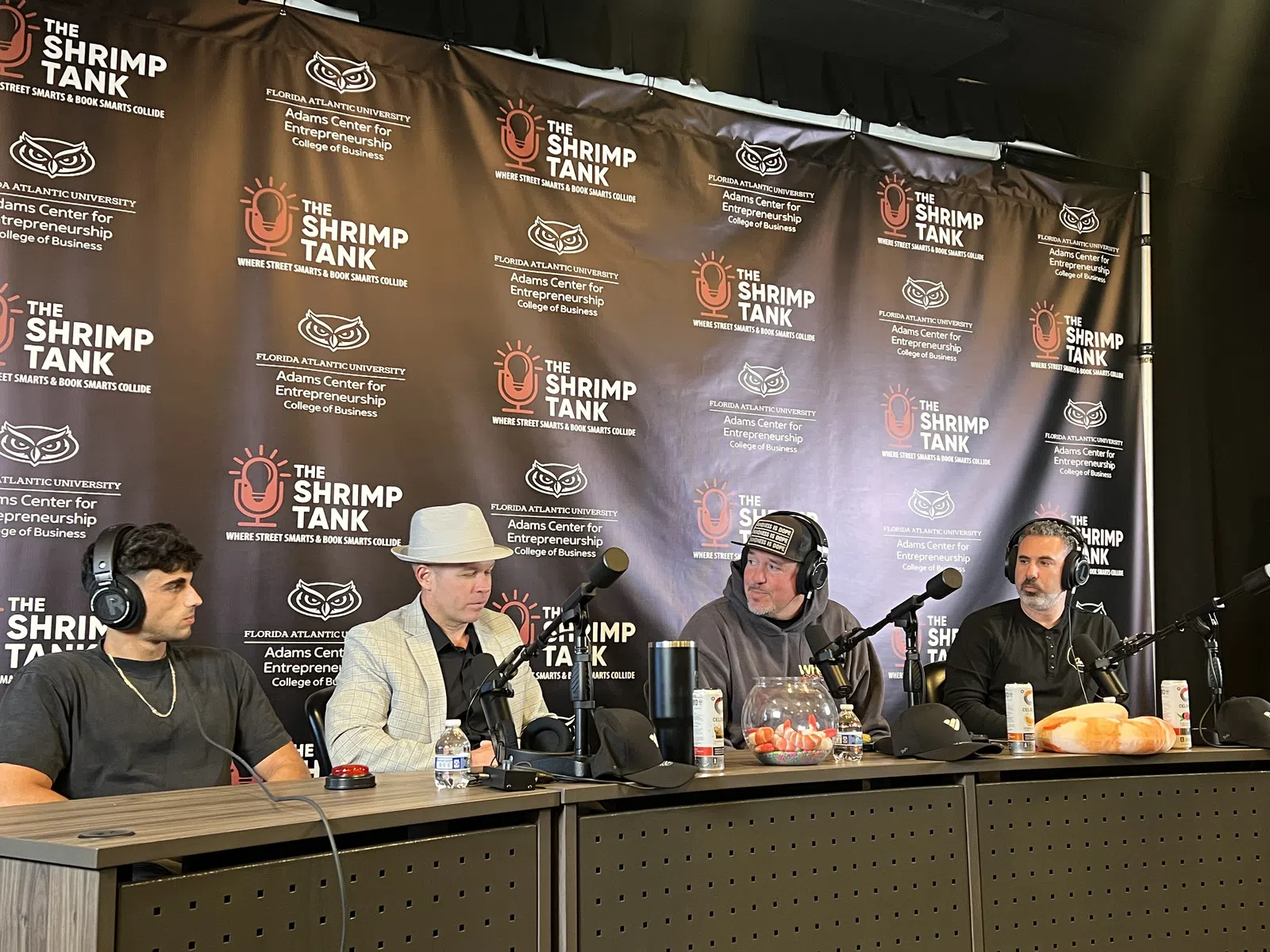 Four men seated at podcast table with microphones under The Shrimp Tank backdrop at Florida Atlantic University event.
