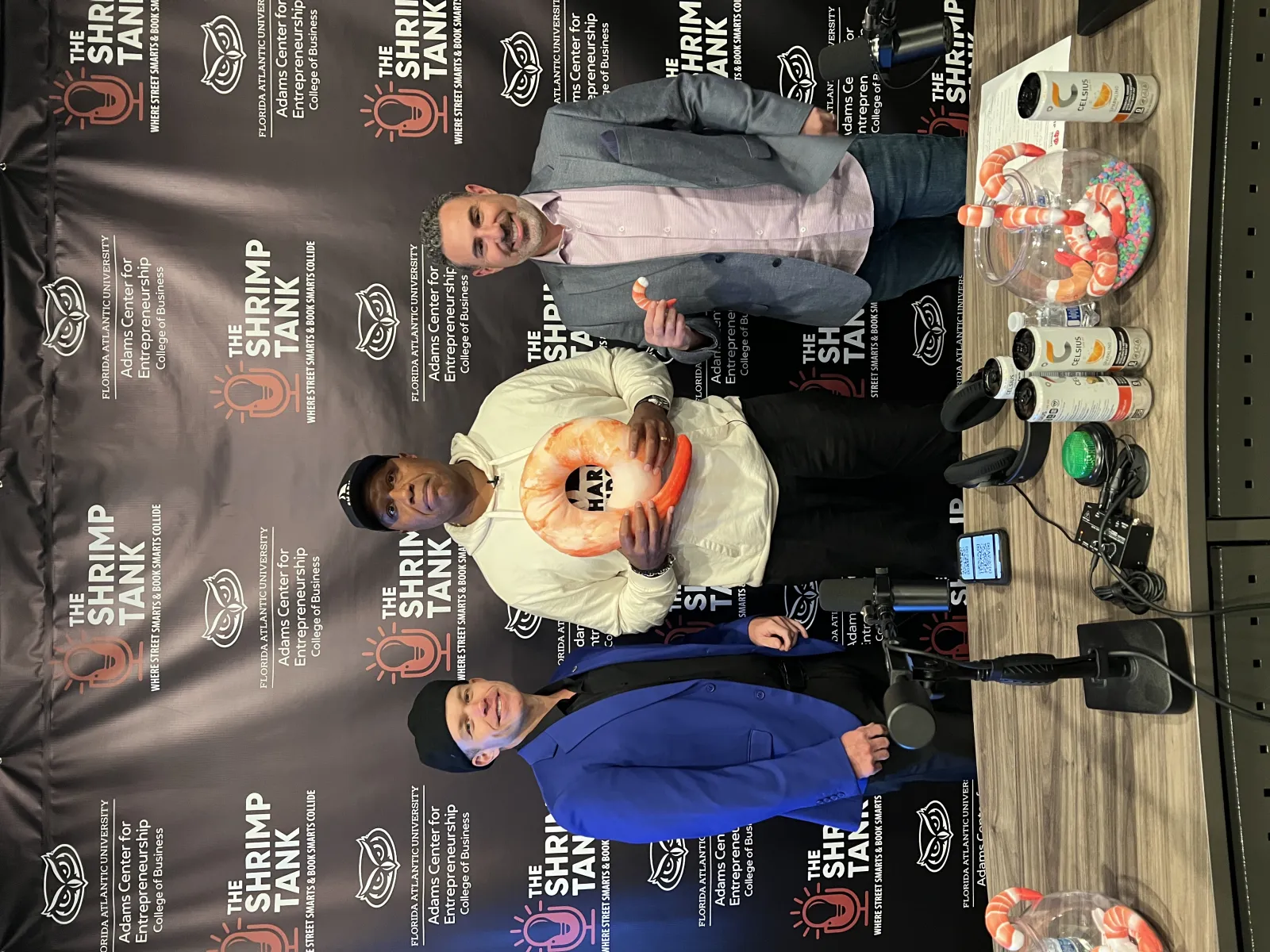 Three men standing at The Shrimp Tank event with shrimp-themed products and microphone on table