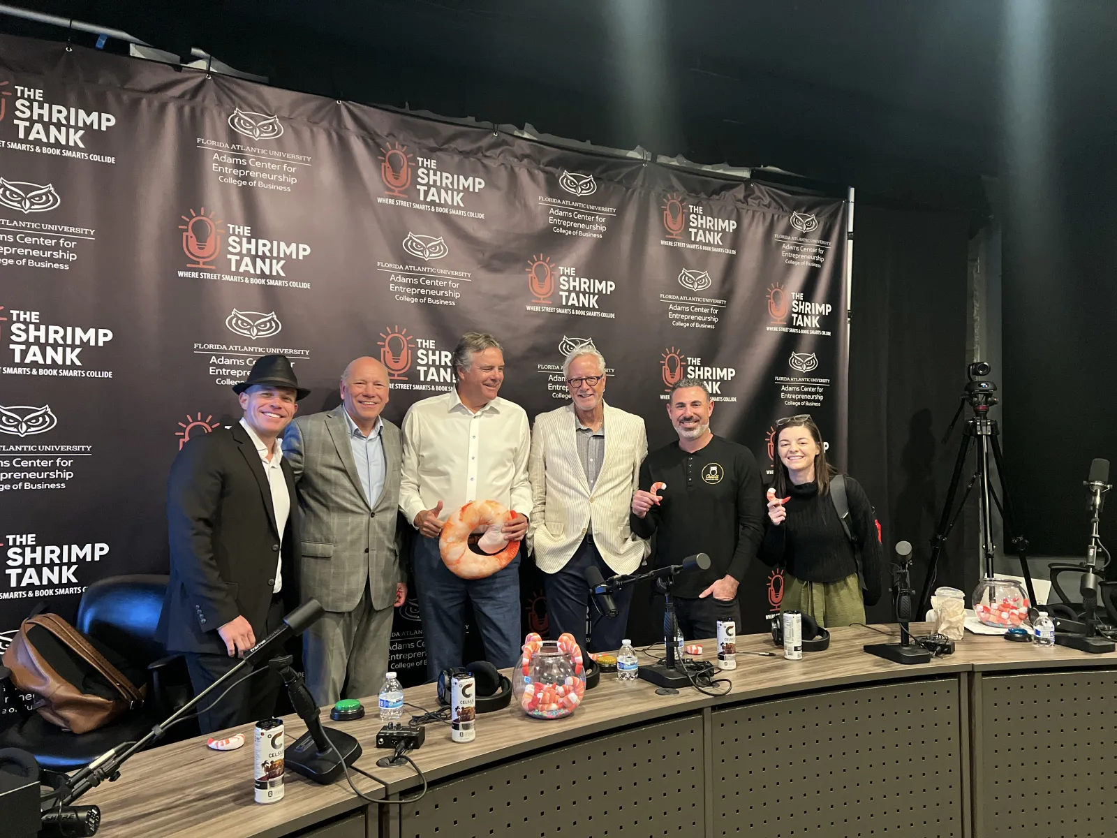 Group of six people posing in front of The Shrimp Tank podcast banner in a recording studio with microphones and props.