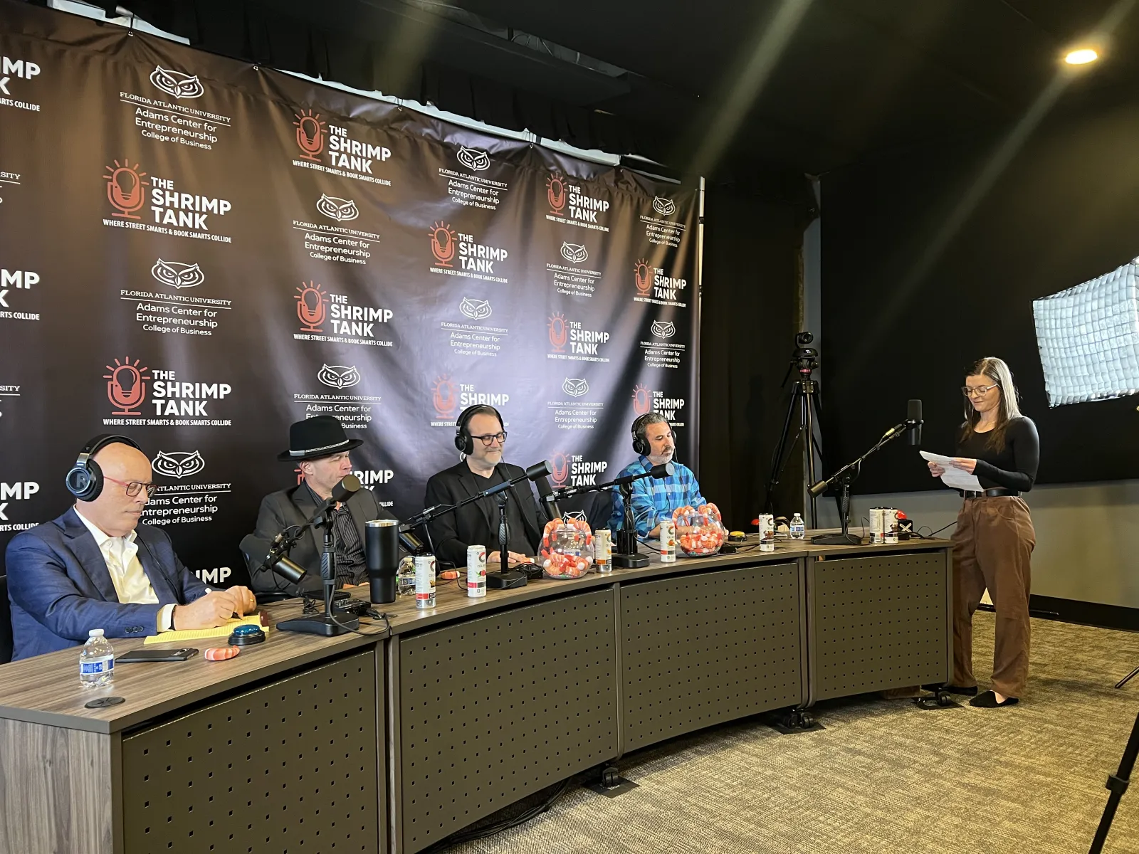 Four judges seated at a panel with microphones and headsets, a woman standing with notes in a podcast studio setup.