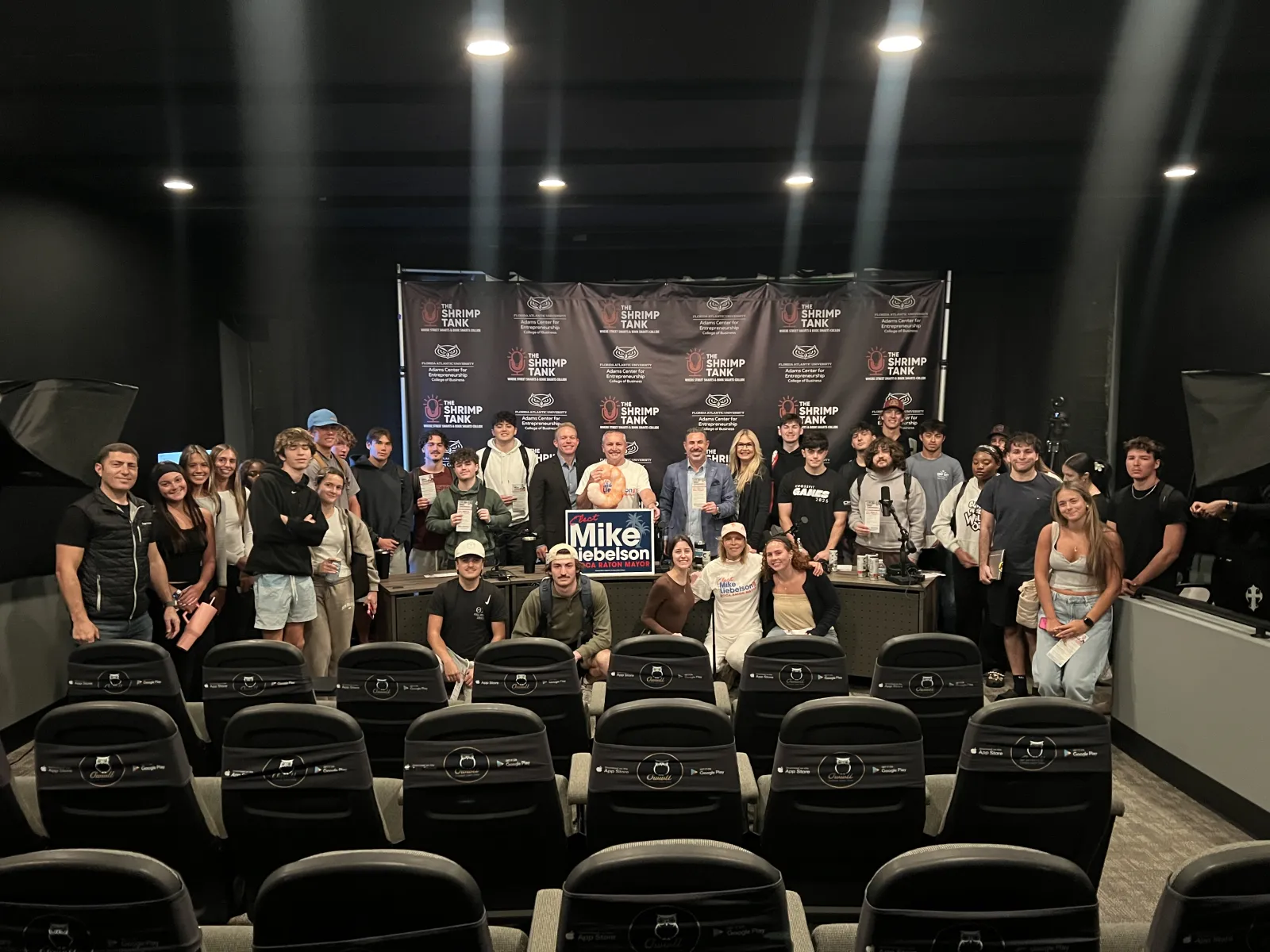 Large group of people posing for a photo in a black room with theater seats and a Mike Nebelson campaign sign