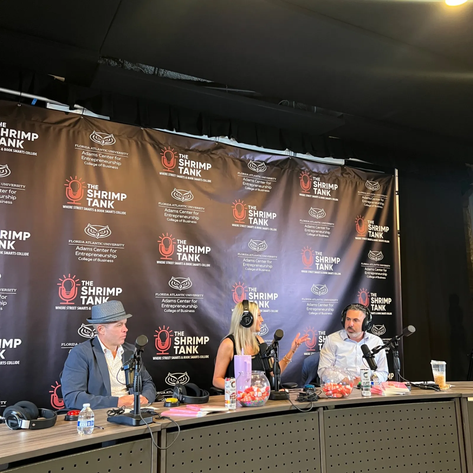 Three people recording a podcast at The Shrimp Tank event with microphones and branded backdrop.