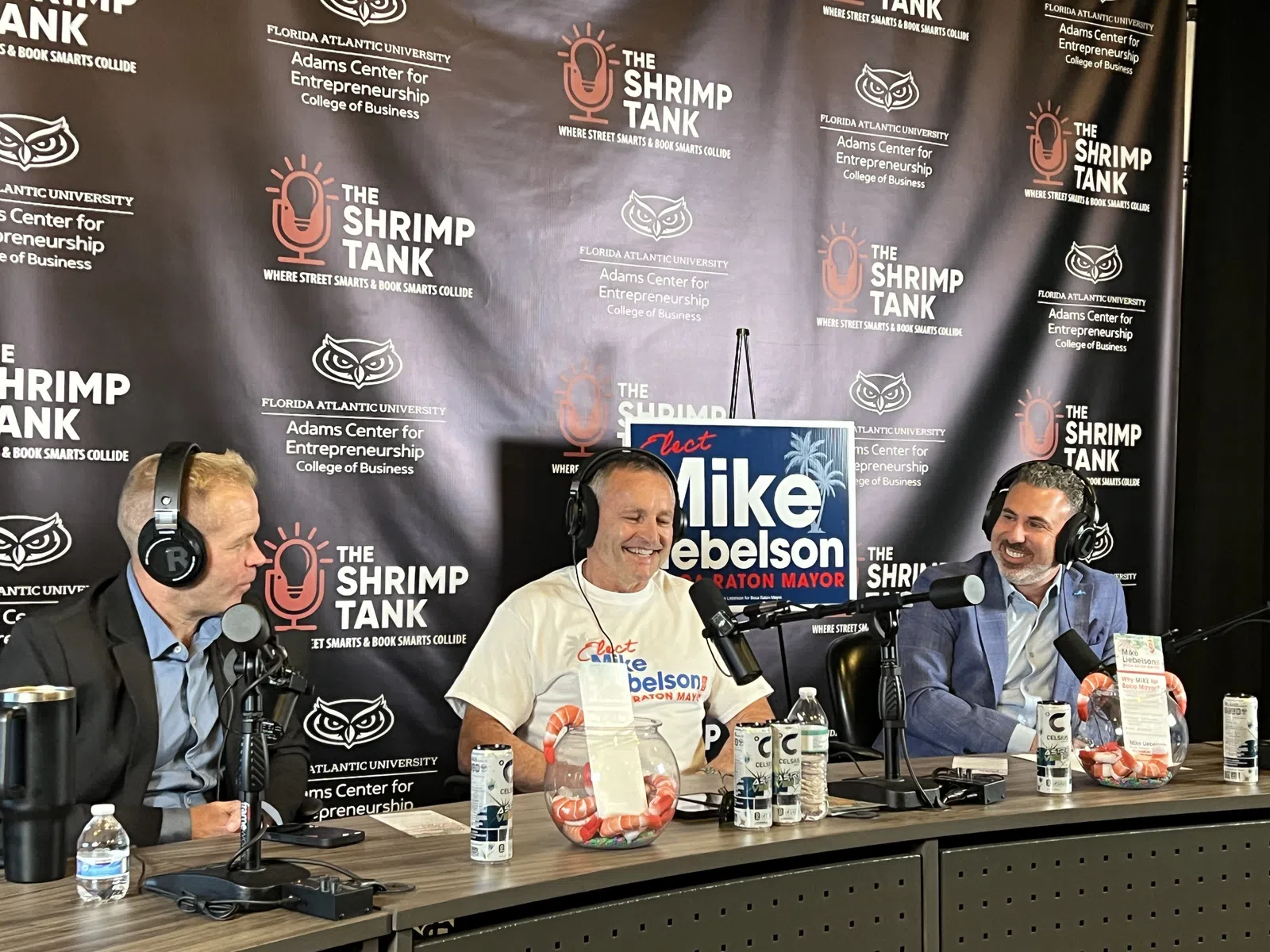 Three men podcasting at The Shrimp Tank with campaign signs and branded backdrop for Mike Debelson for mayor.