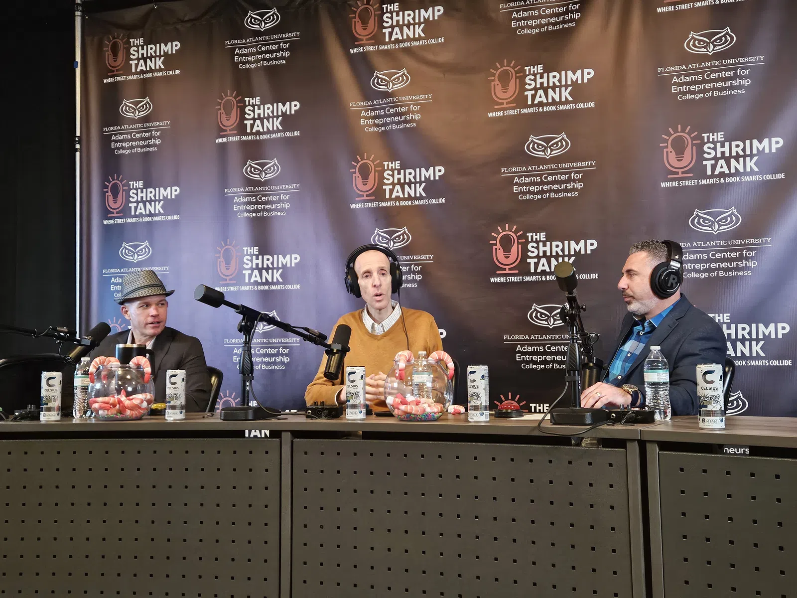 Three men wearing headphones sit at a table with microphones in front of a backdrop promoting The Shrimp Tank podcast.