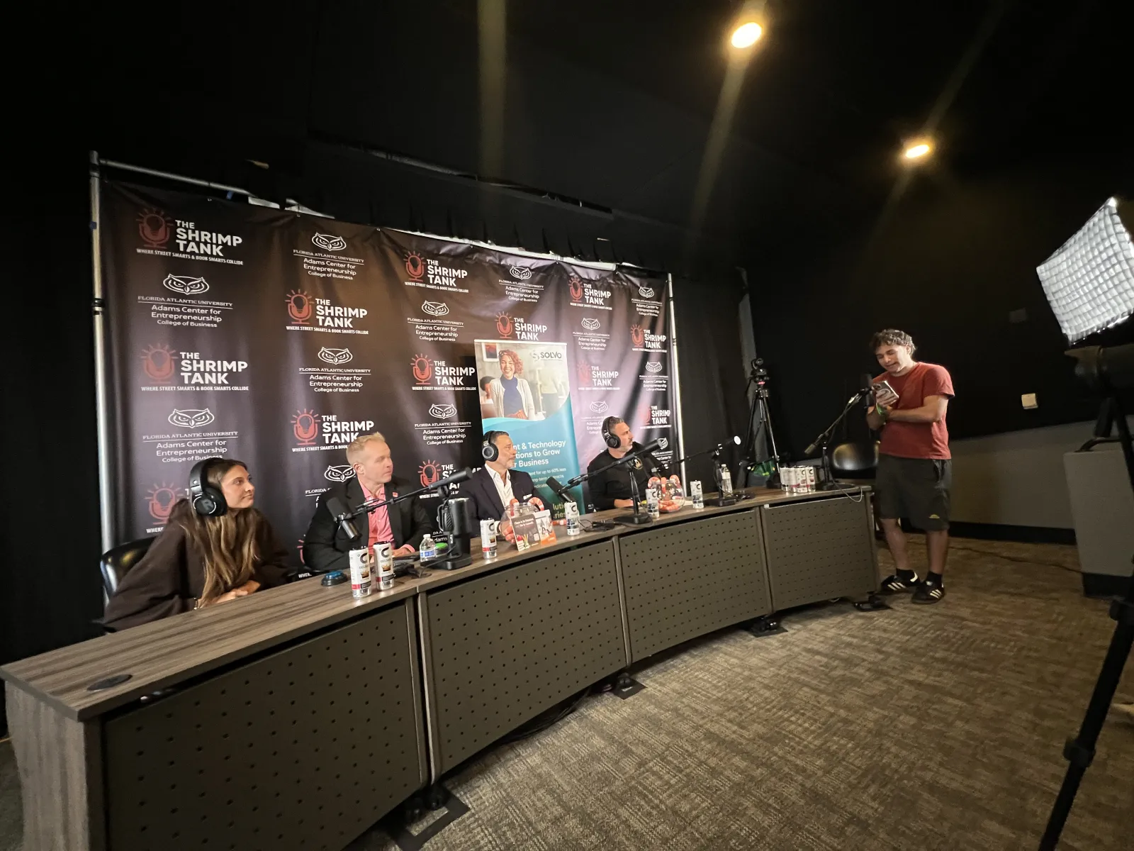 Four hosts wearing headphones talk behind a curved desk with Shrimp Tank banners and a cameraman filming them.