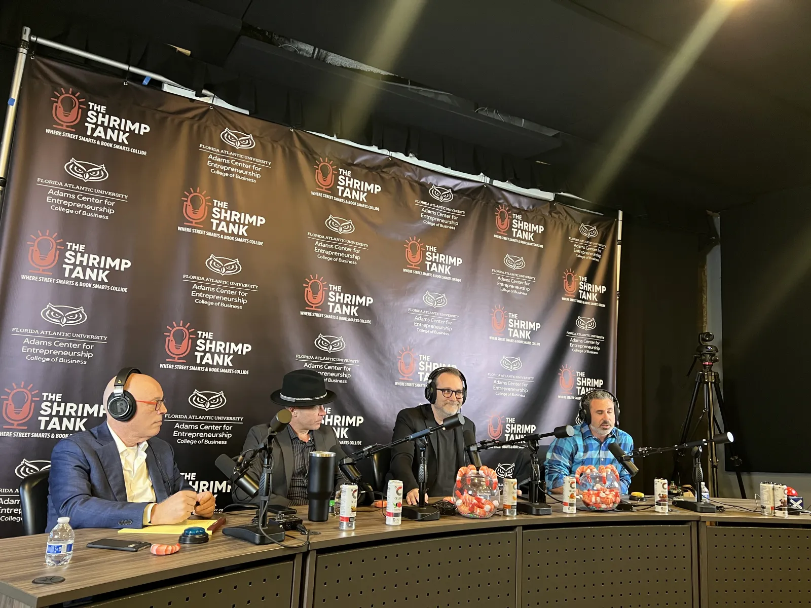 Four men with headsets sitting at a podcast table with microphones and The Shrimp Tank backdrop