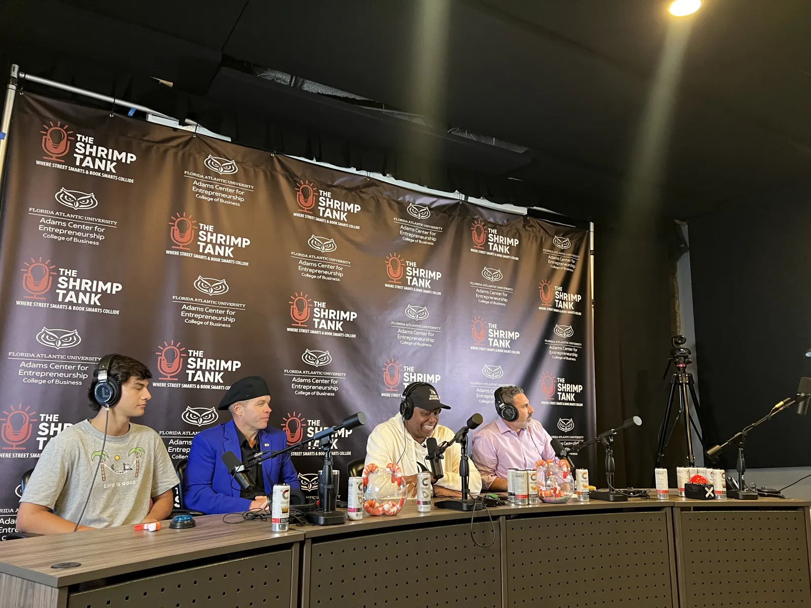 Four men with headphones sitting at a podcast panel table with microphones and branded backdrop of The Shrimp Tank event