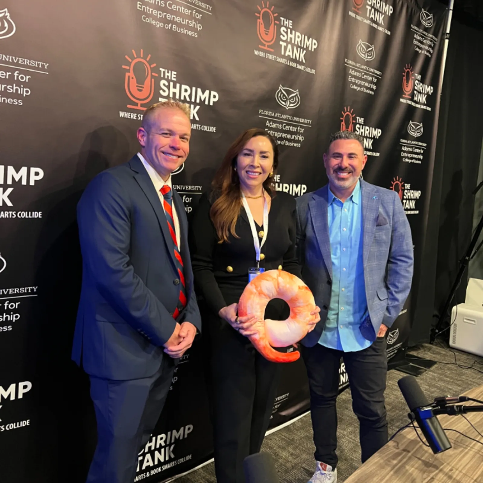 Three professionals posing at The Shrimp Tank event with branded backdrop and shrimp-shaped award