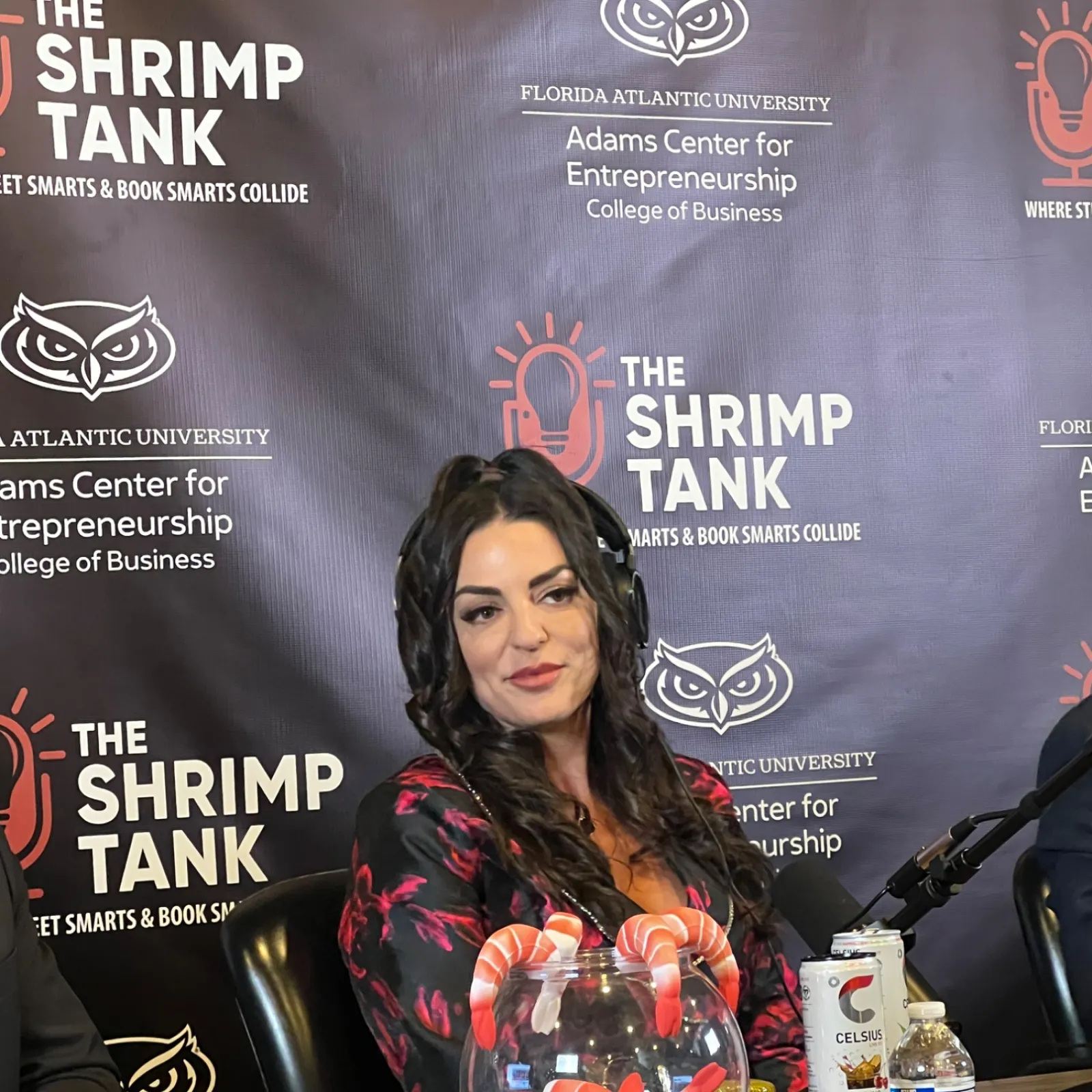 Woman sitting at desk with bowl of shrimp, microphone, and branded backdrop of The Shrimp Tank entrepreneurship event.