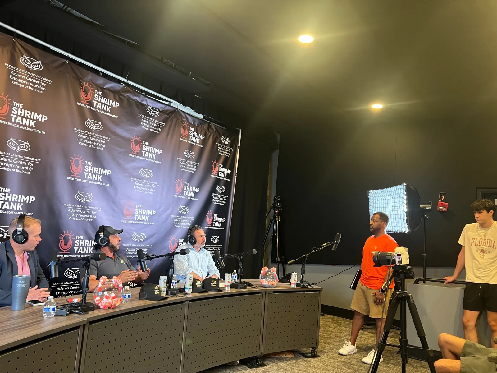 Podcast recording with four hosts behind microphones and two people standing near a camera and light setup.