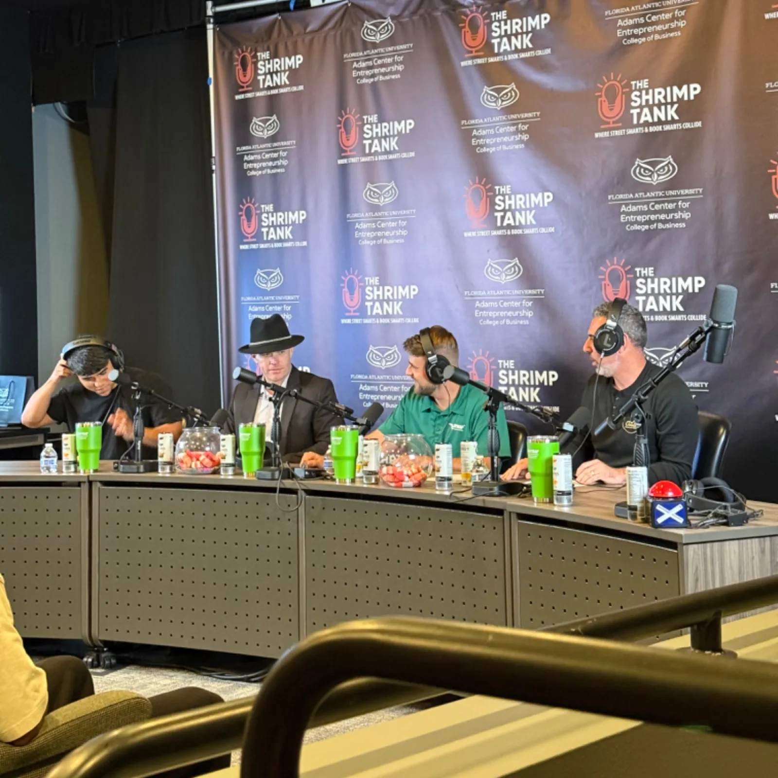 Four people wearing headphones at a podcast table with The Shrimp Tank banner and microphones