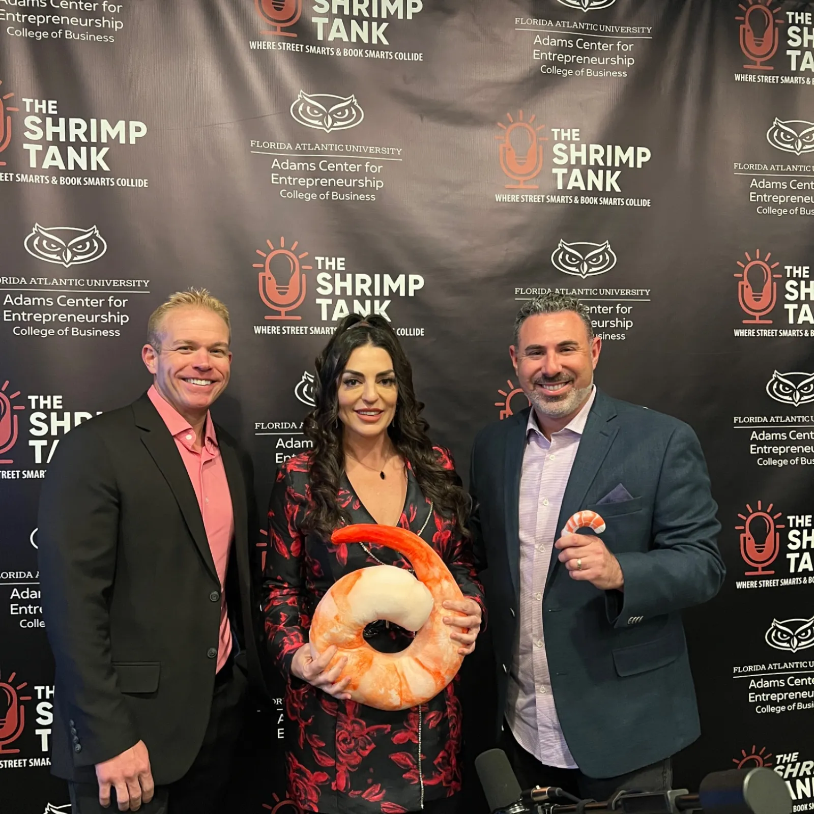 Three people pose smiling in front of a Shrimp Tank branded backdrop, one holding a large shrimp-shaped item.