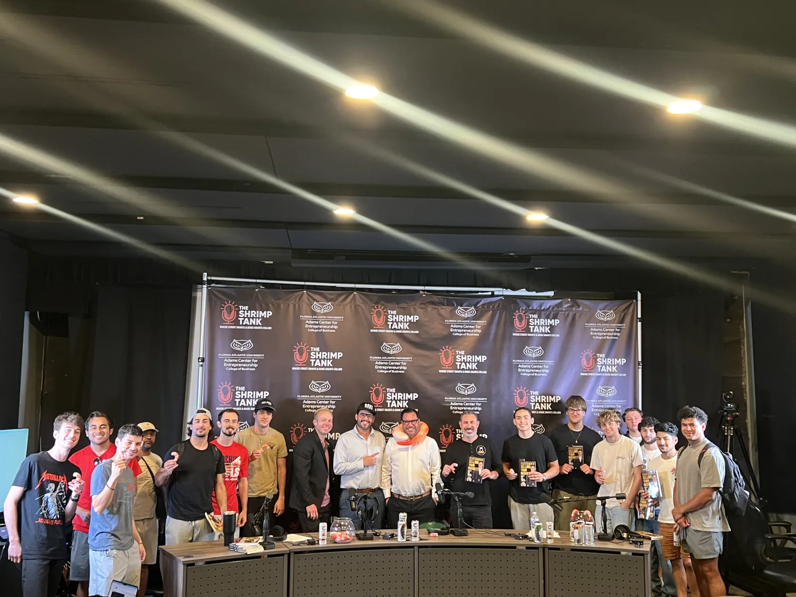 Group photo of smiling people on stage at The Shrimp Tank event with backdrop and trophies.