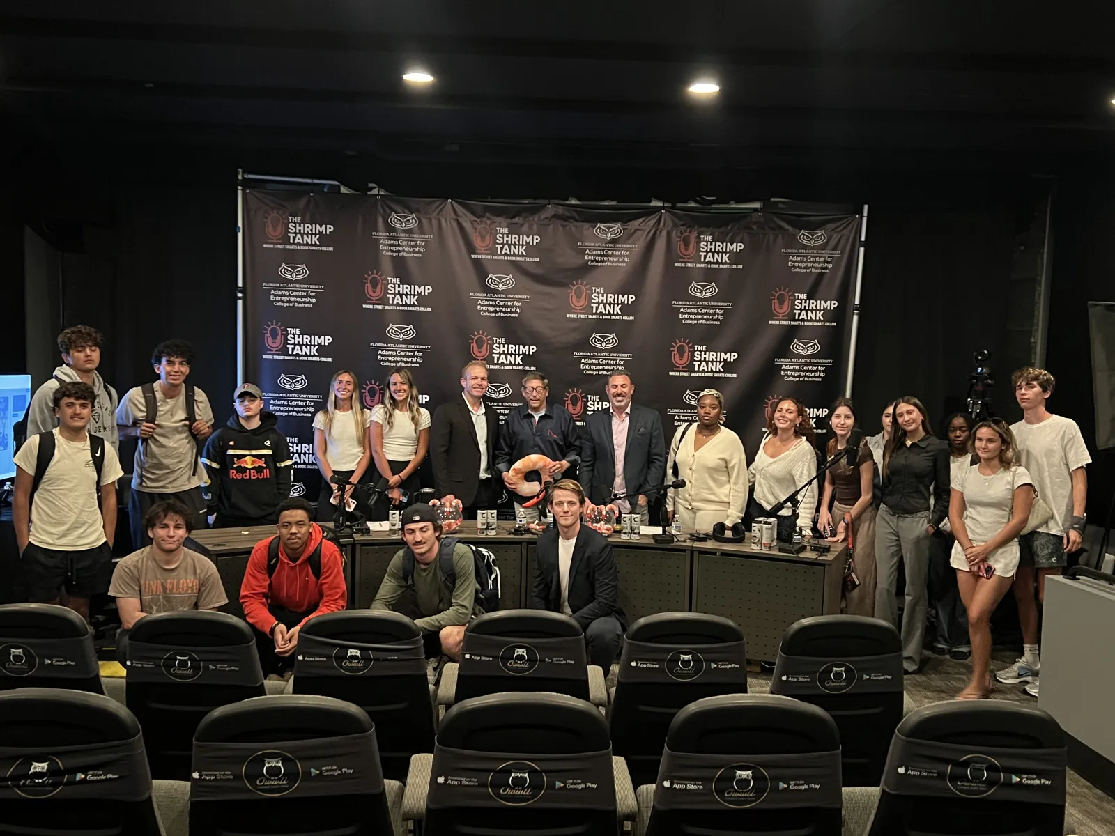 Group photo of diverse people standing and sitting in front of The Shrimp Tank branded backdrop in an event setting.