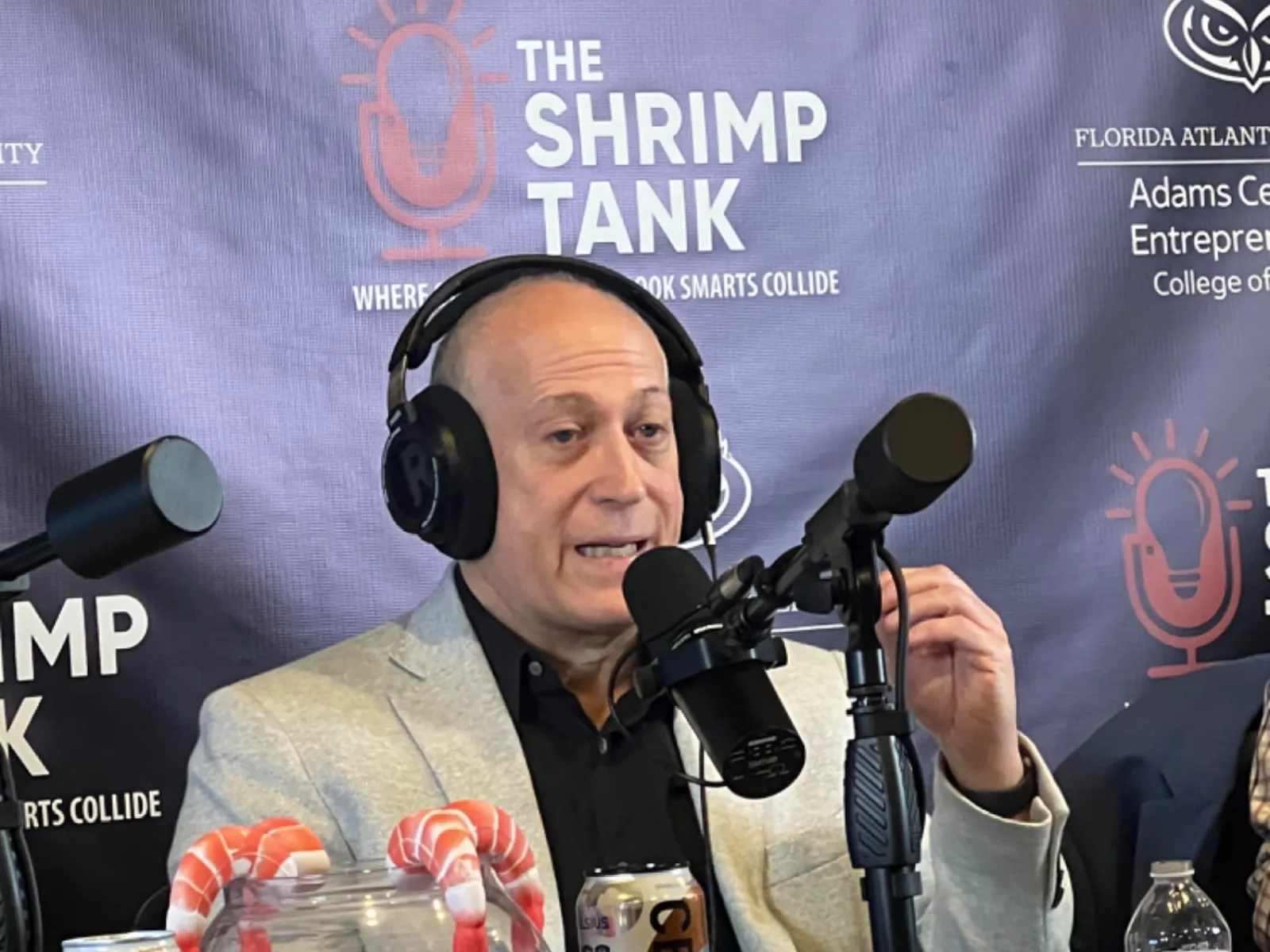 Man wearing headphones speaking into microphone during The Shrimp Tank podcast with shrimp candy jar and Celsius drink cans on desk