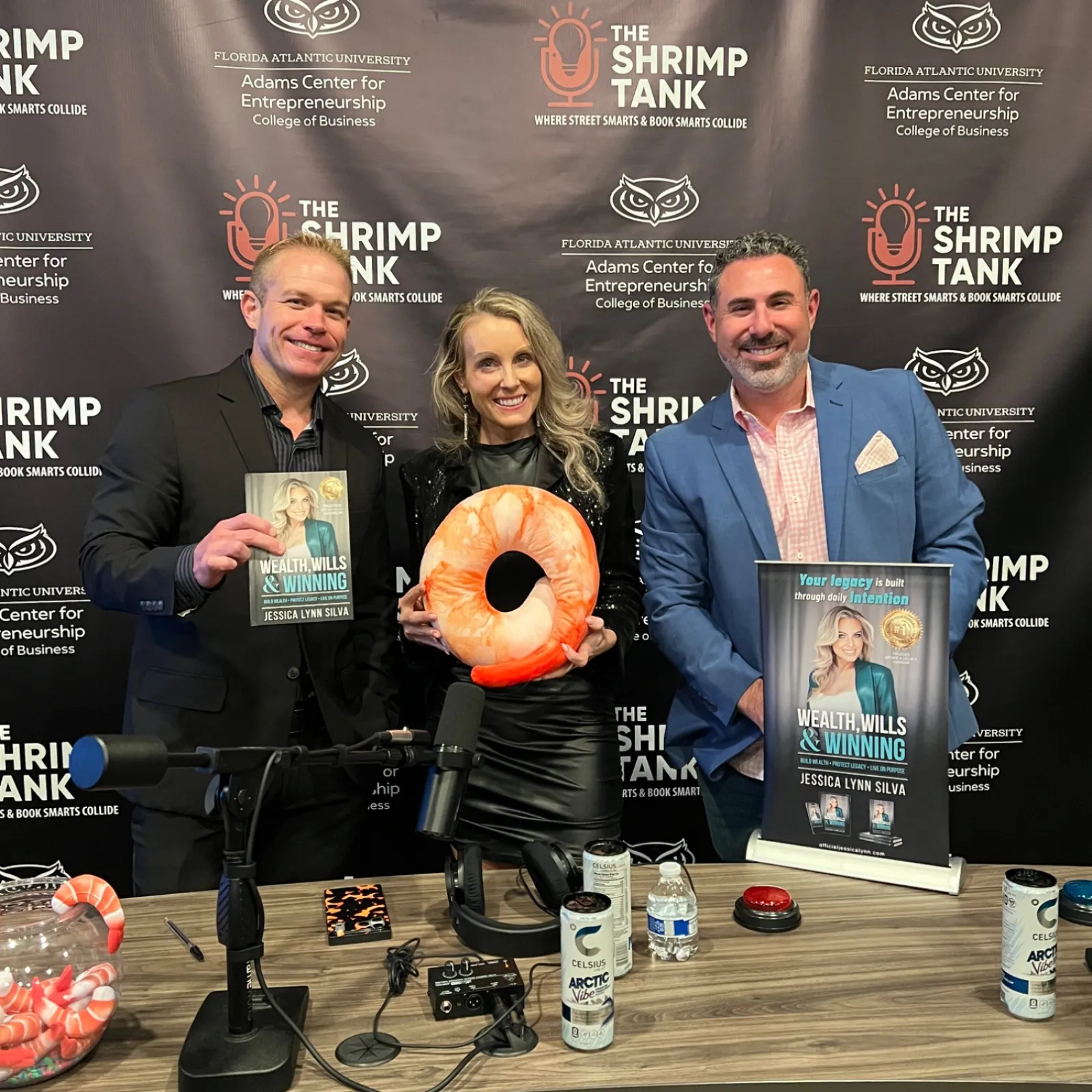 Three people at a podcast table with microphones, holding books and a shrimp-shaped cushion, Shrimp Tank backdrop.