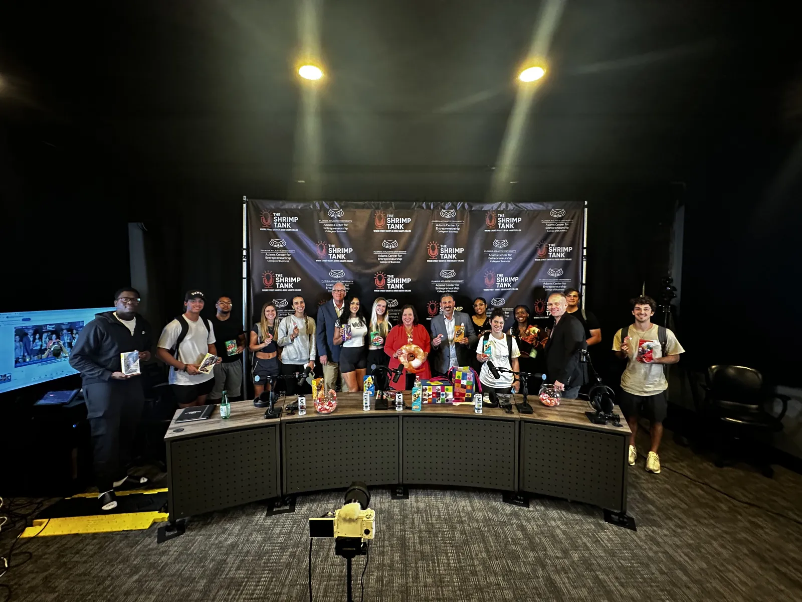 Group photo of attendees at a media event, featuring a branded backdrop and diverse individuals holding merchandise.