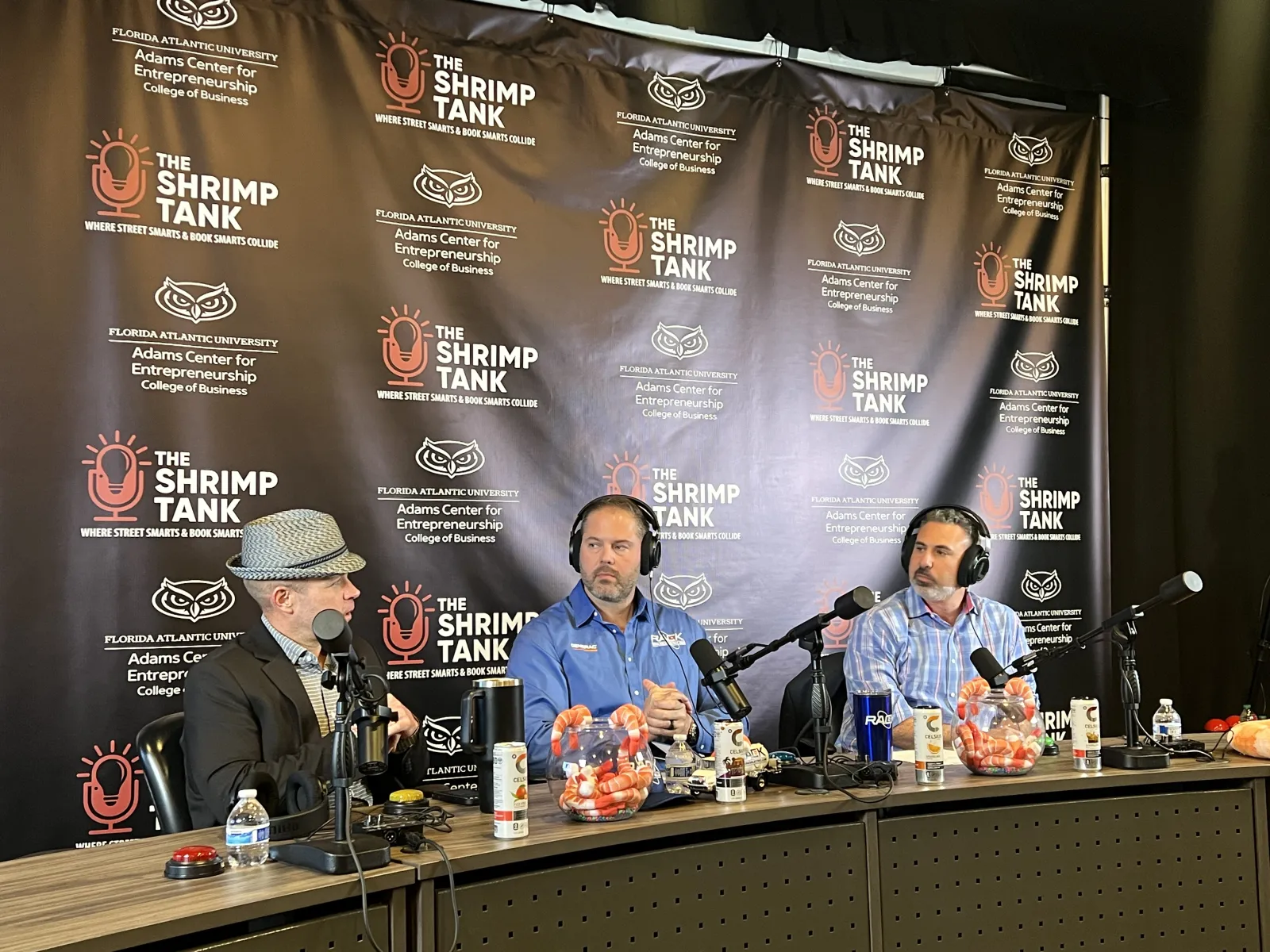 Three men with microphones hosting a podcast in front of The Shrimp Tank backdrop at Florida Atlantic University.