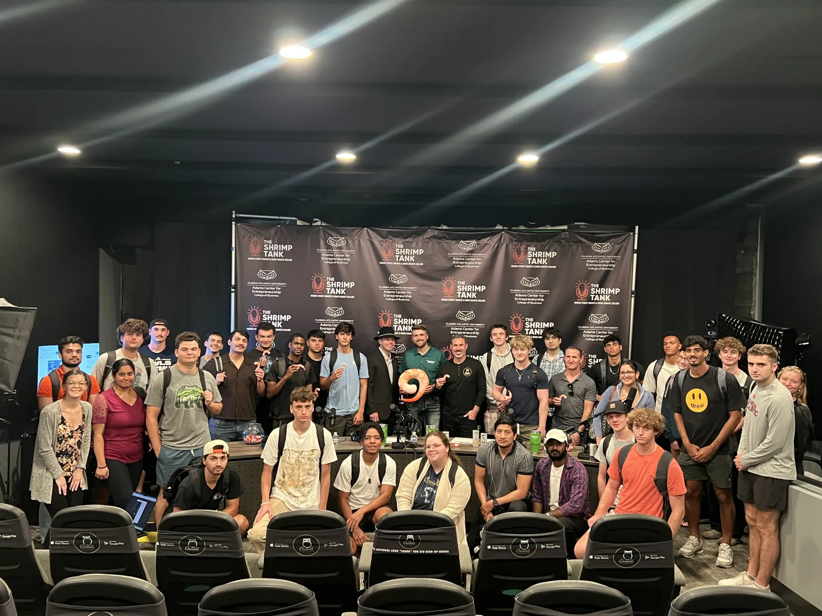 Large diverse group posing in front of a Shrimp Tank banner in an indoor event space with studio lighting.