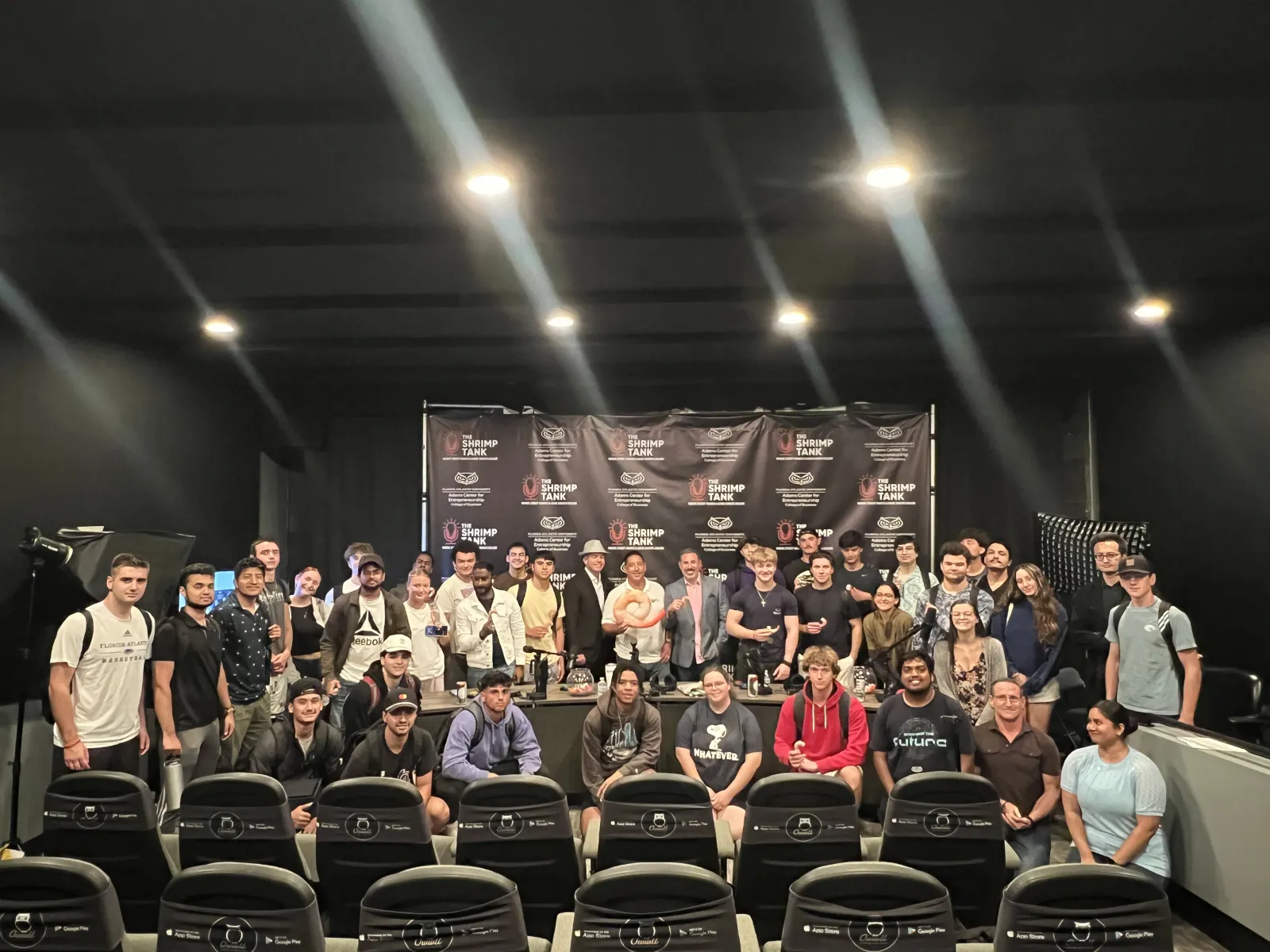 Group photo of diverse people posing in front of a Shrimp Tank event backdrop with studio lighting.