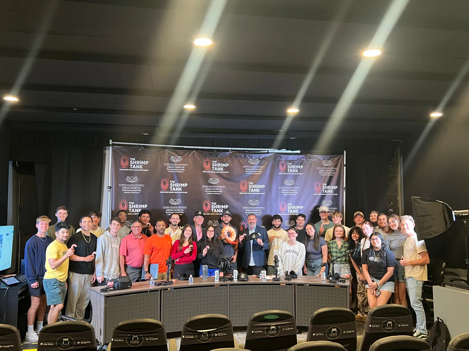 Large group of people posing in front of The Shrimp Tank backdrop with studio lighting and microphones.