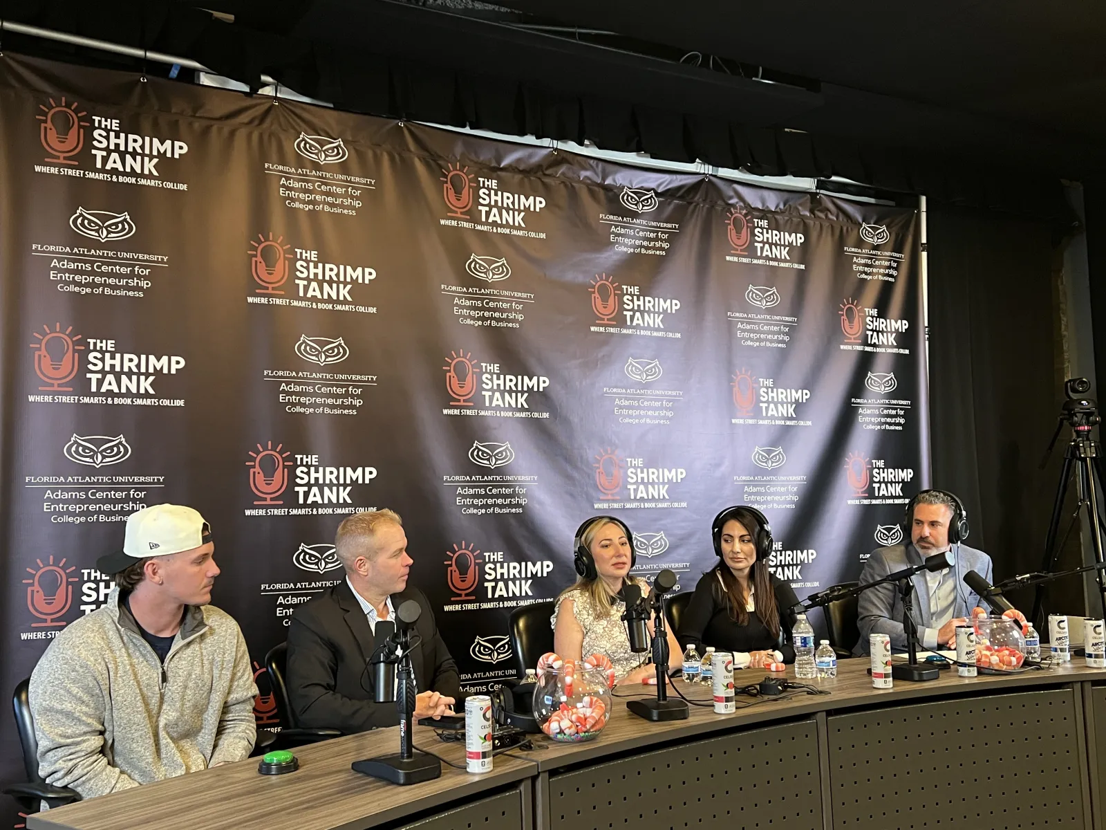 Panel of five speakers with microphones at The Shrimp Tank event backdrop discussing entrepreneurship.