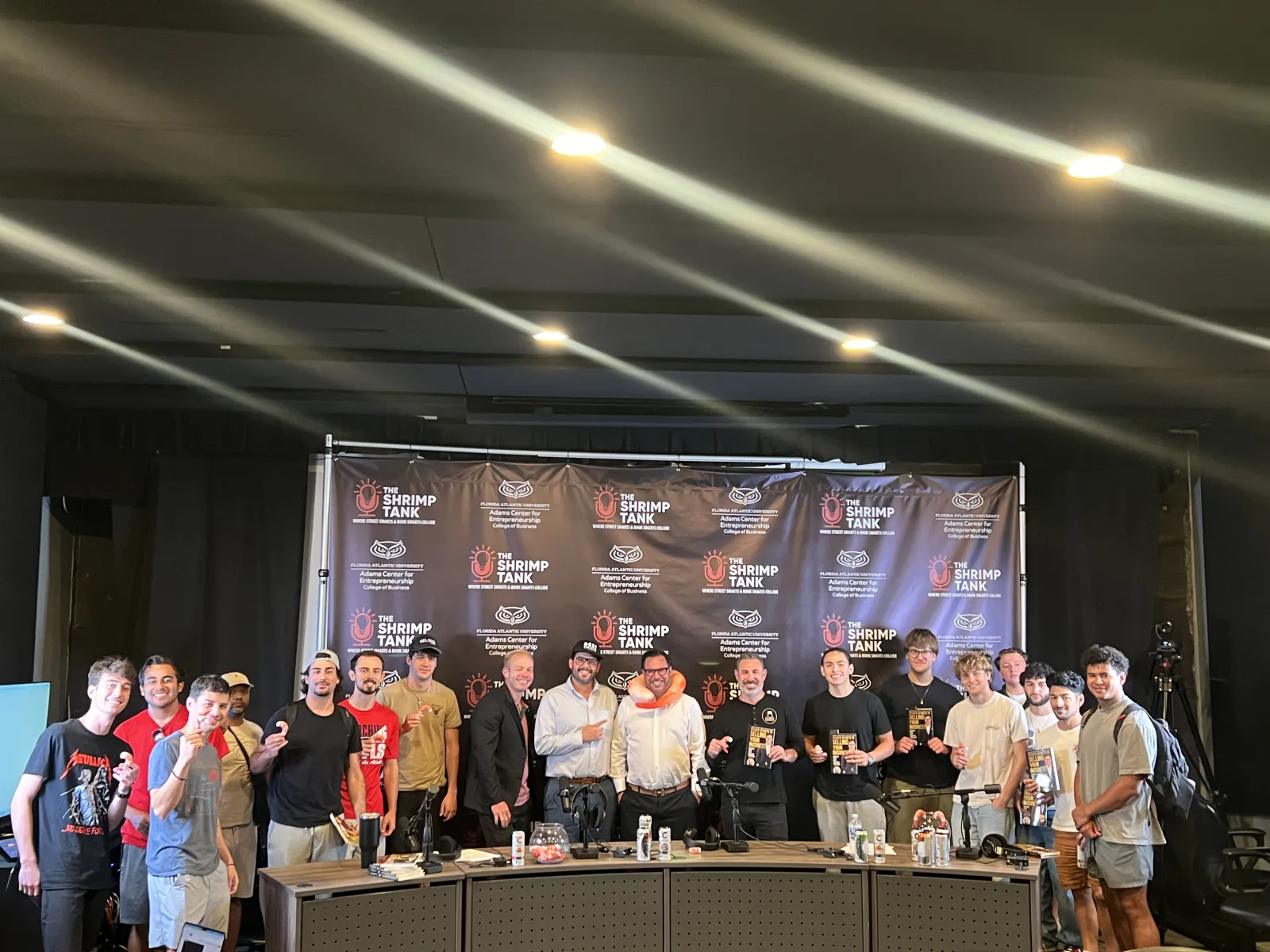 Group of people posing in front of The Shrimp Tank event backdrop on stage with awards and trophies