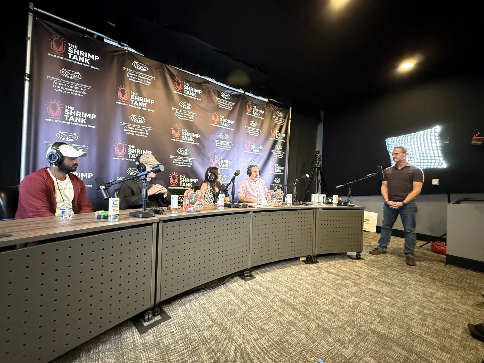 Panel of four judges seated with microphones facing a standing presenter in a podcast studio with The Shrimp Tank backdrop